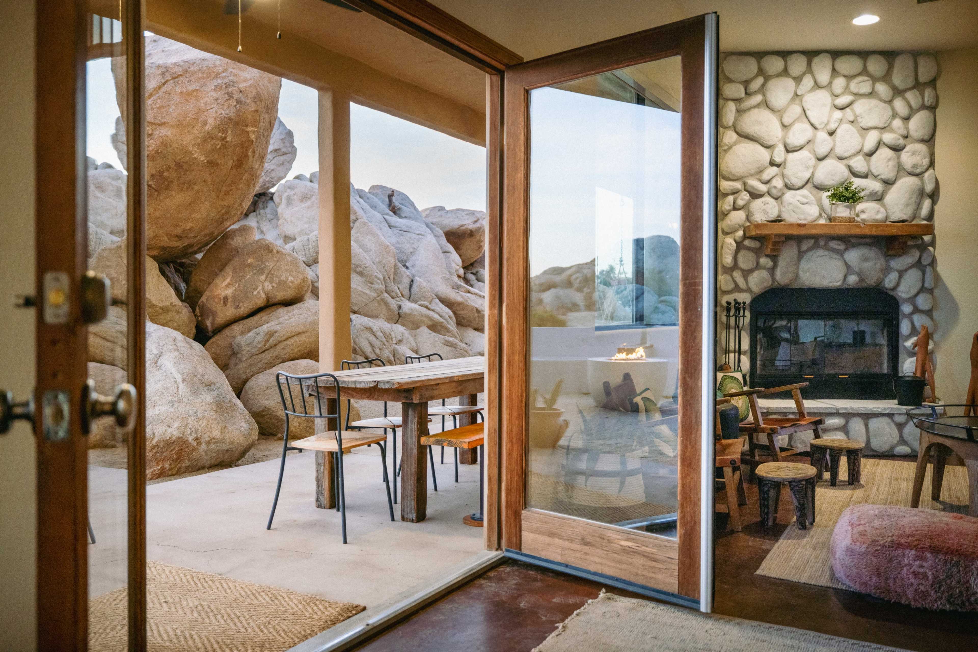 The image shows a spacious interior with wooden doors open to a patio surrounded by large rocks, featuring a dining table and a fireplace.