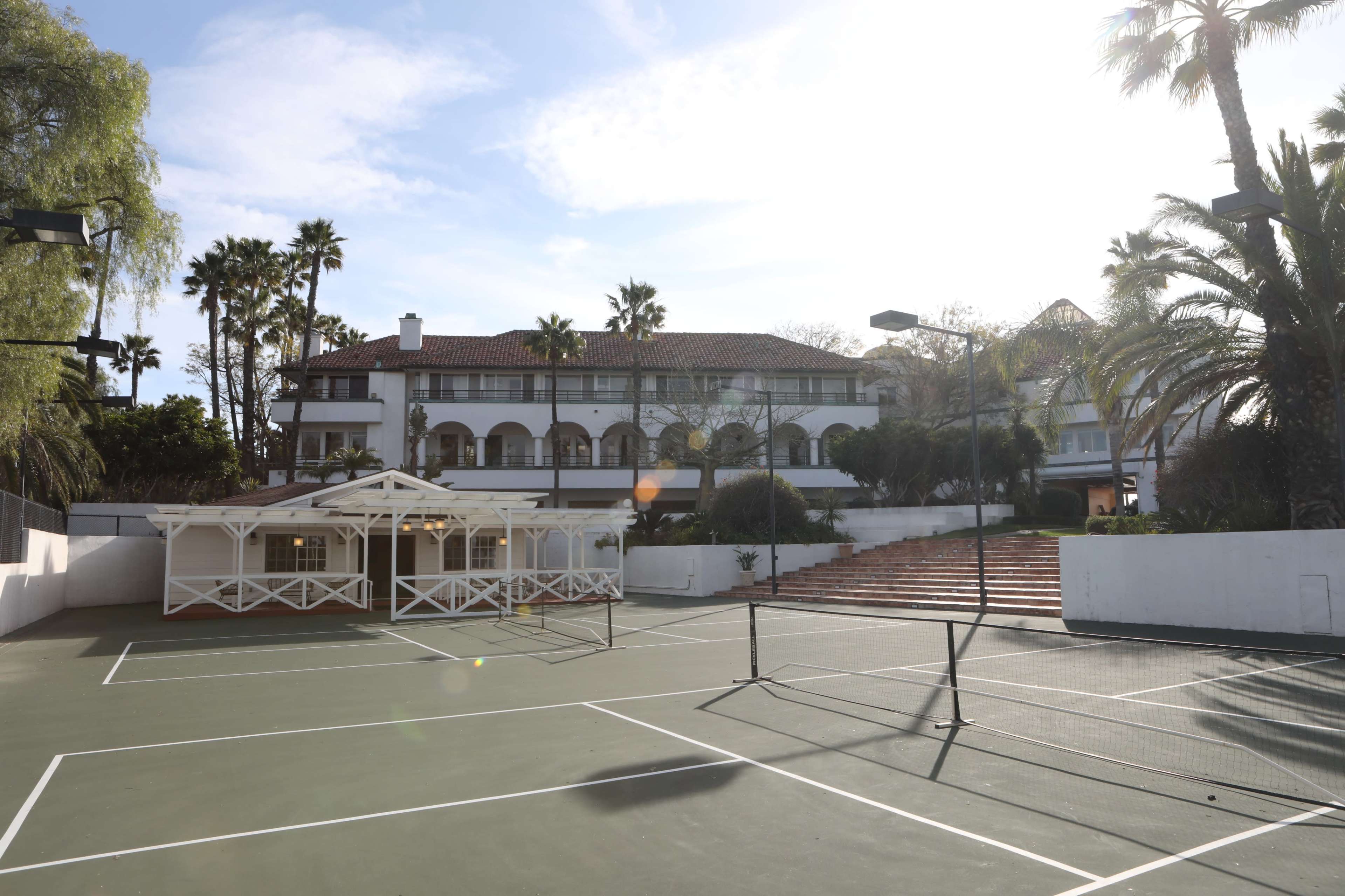 A tennis court with a view of a large building surrounded by palm trees under a bright sky.