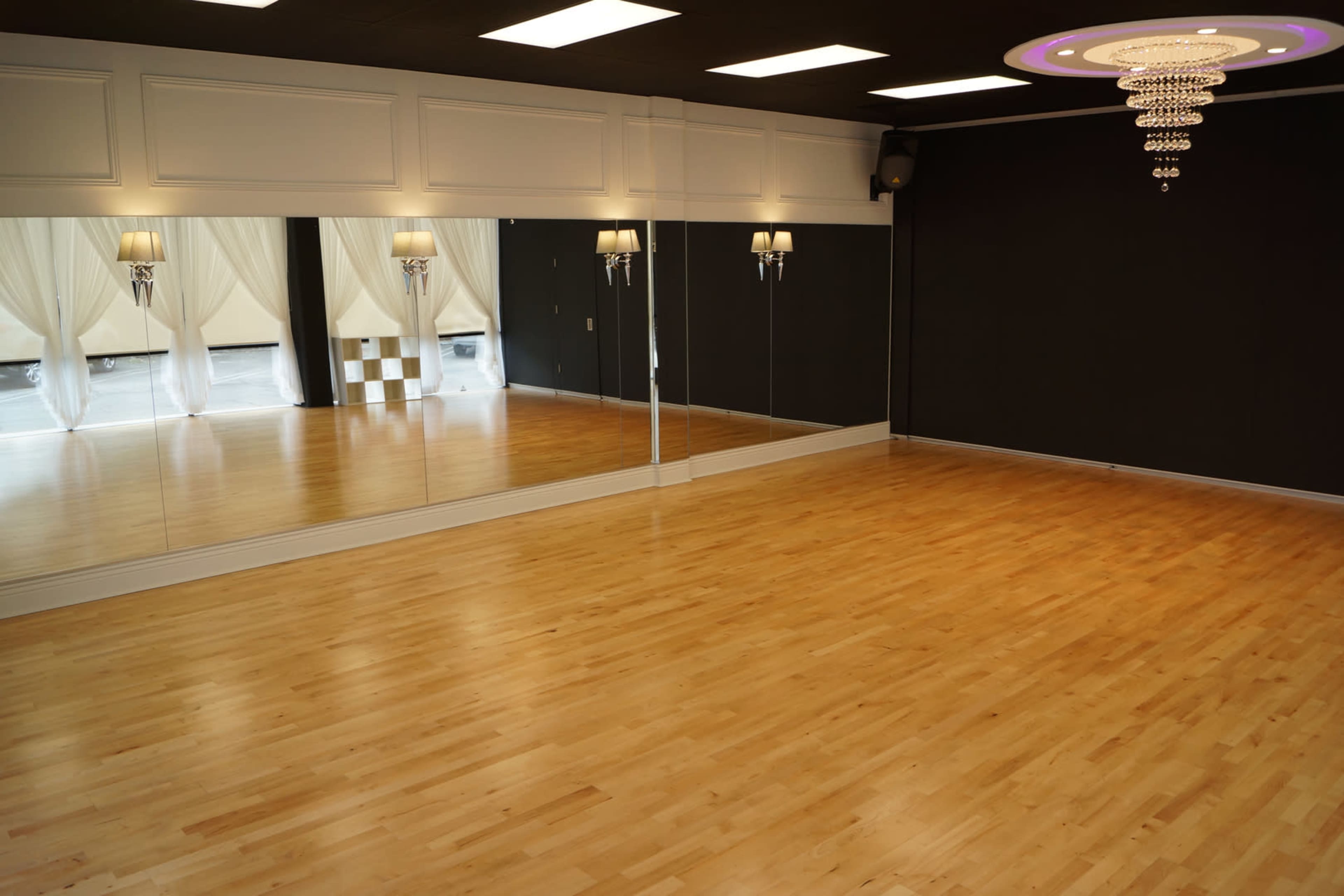 A spacious dance studio featuring wooden flooring, a large mirror on one wall, and a modern chandelier hanging from the ceiling.