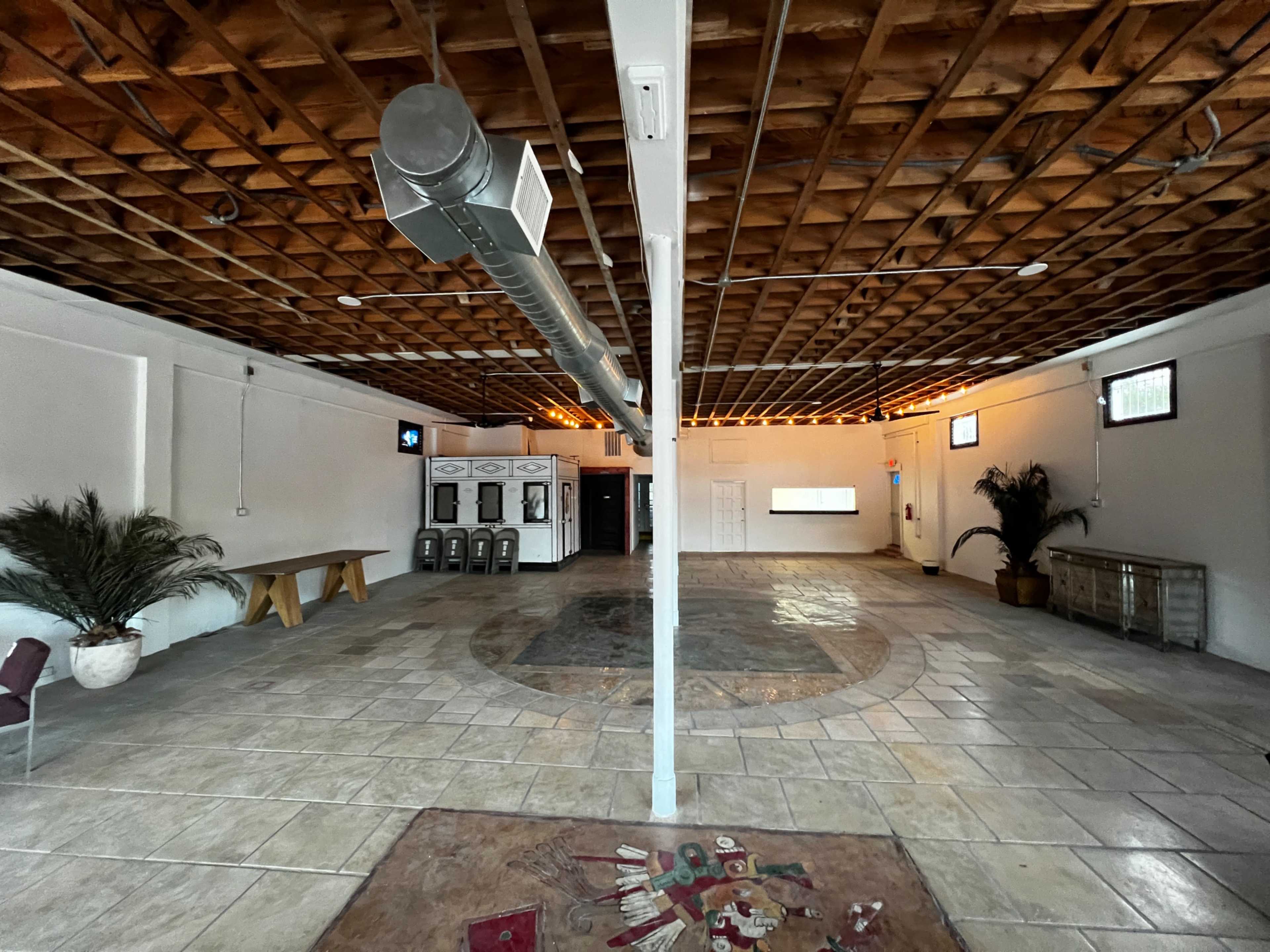 A spacious room with a tiled floor, exposed wooden beams, and a central circular design, featuring a few decorative plants and minimal furnishings.