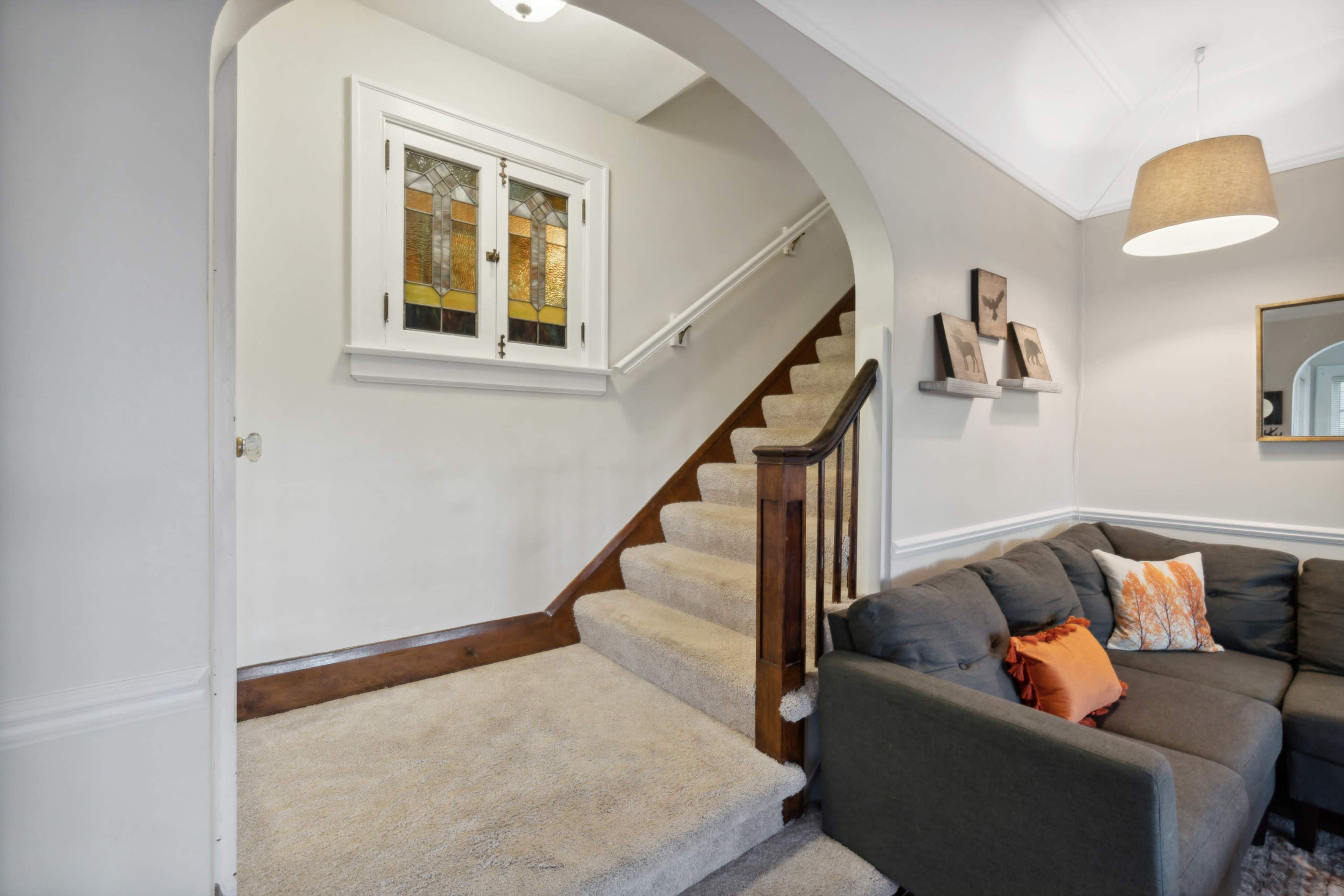 A stairway with a stained-glass window leads to an upper floor in a living room featuring a gray sectional sofa and decorative pillows.