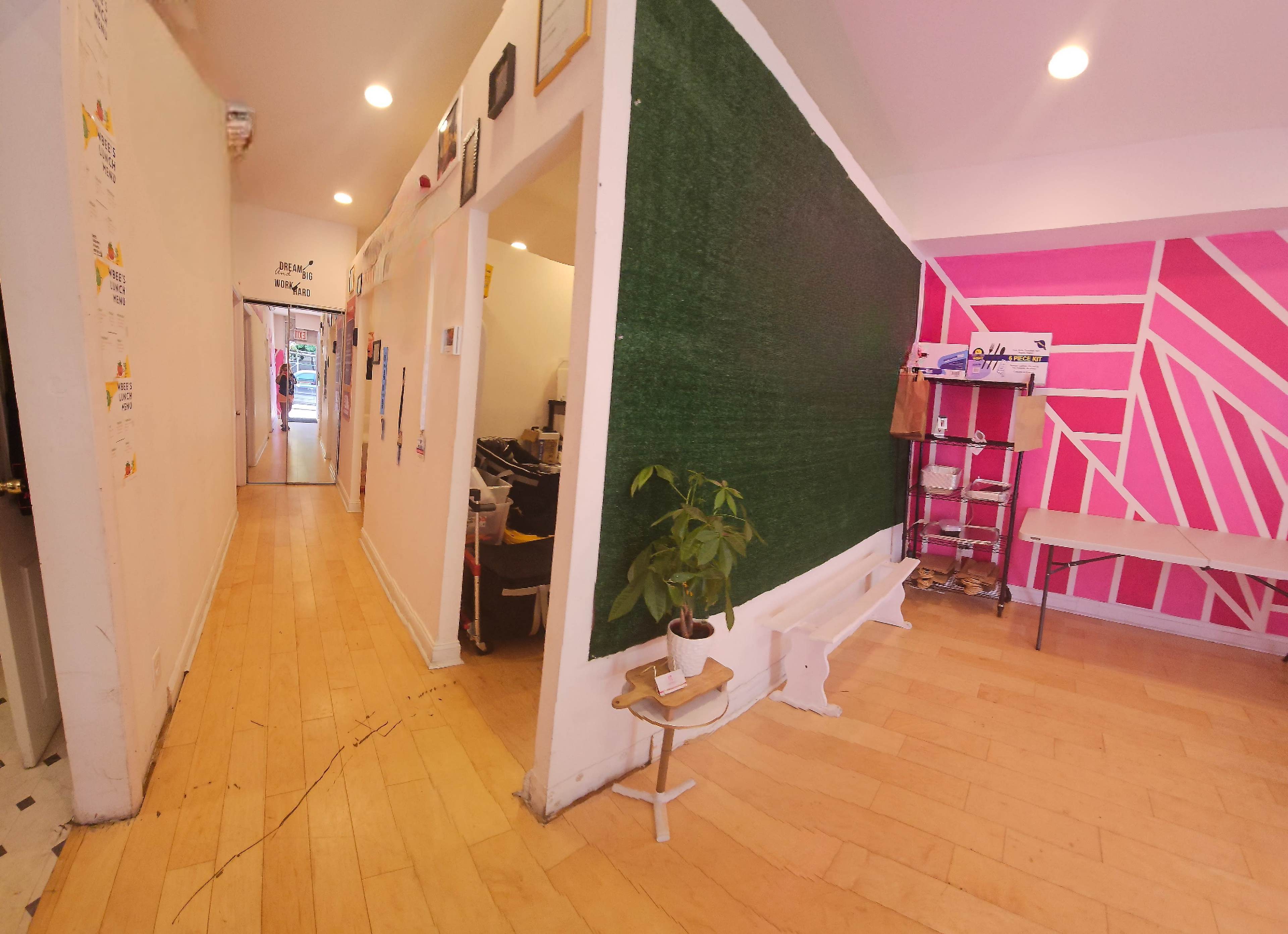 A bright interior space with a green wall on one side and pink geometric patterns on another, featuring wooden flooring and a small plant on a pedestal.
