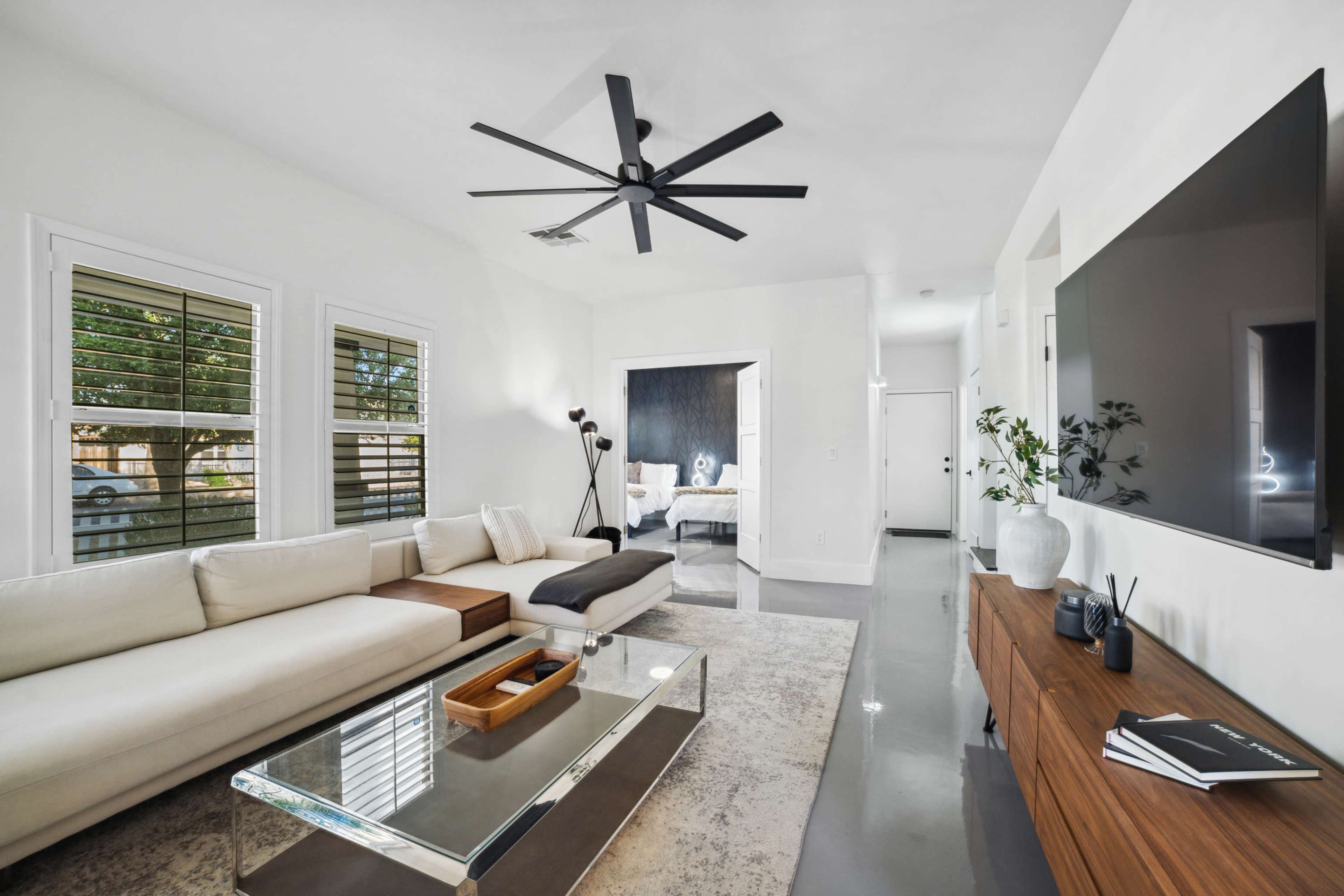 A spacious living room features a sectional sofa, a glass coffee table, a large wall-mounted television, and minimalist decor with natural light from the windows.