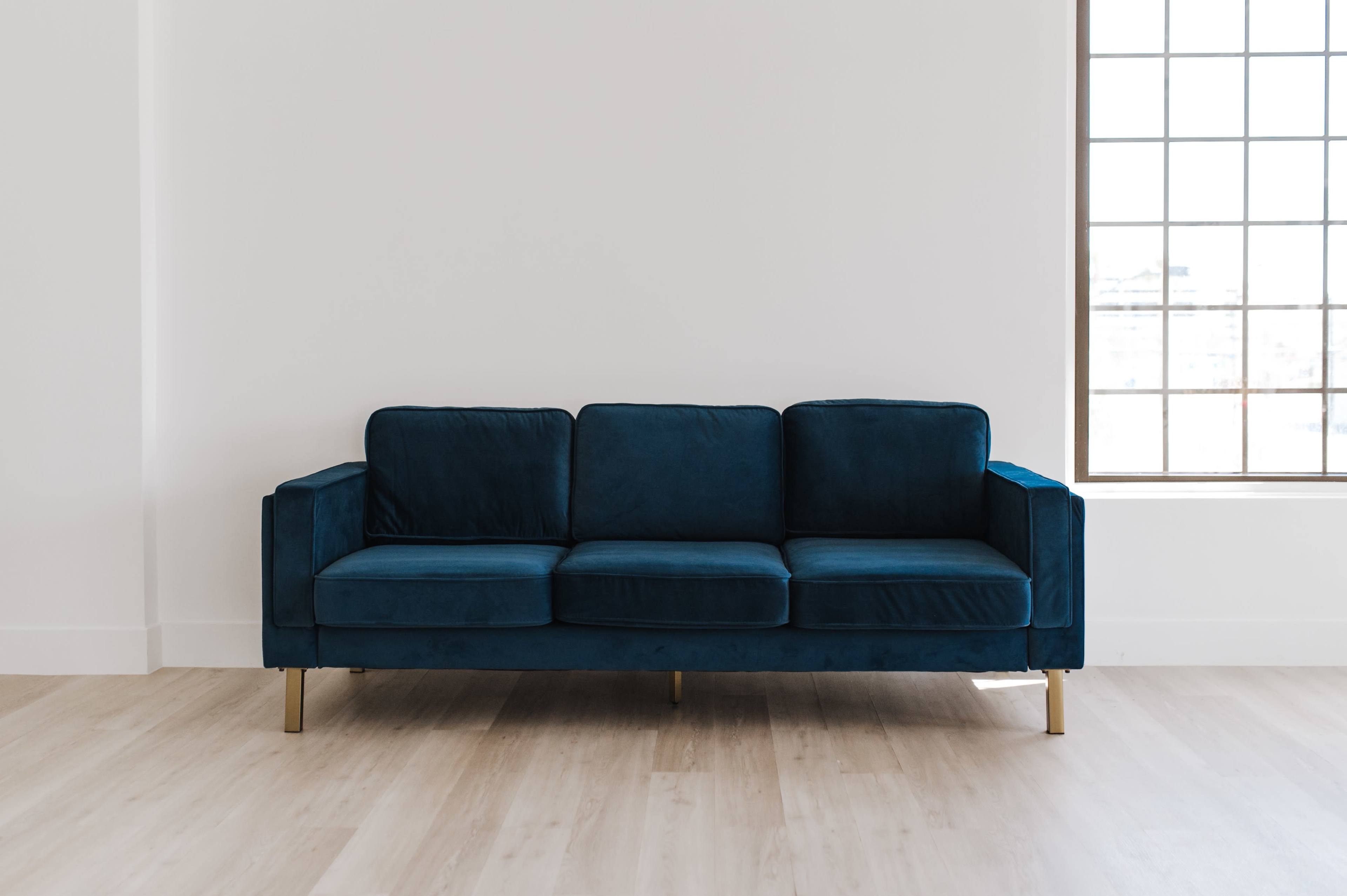 A blue velvet sofa with gold legs is positioned against a white wall next to a large window.