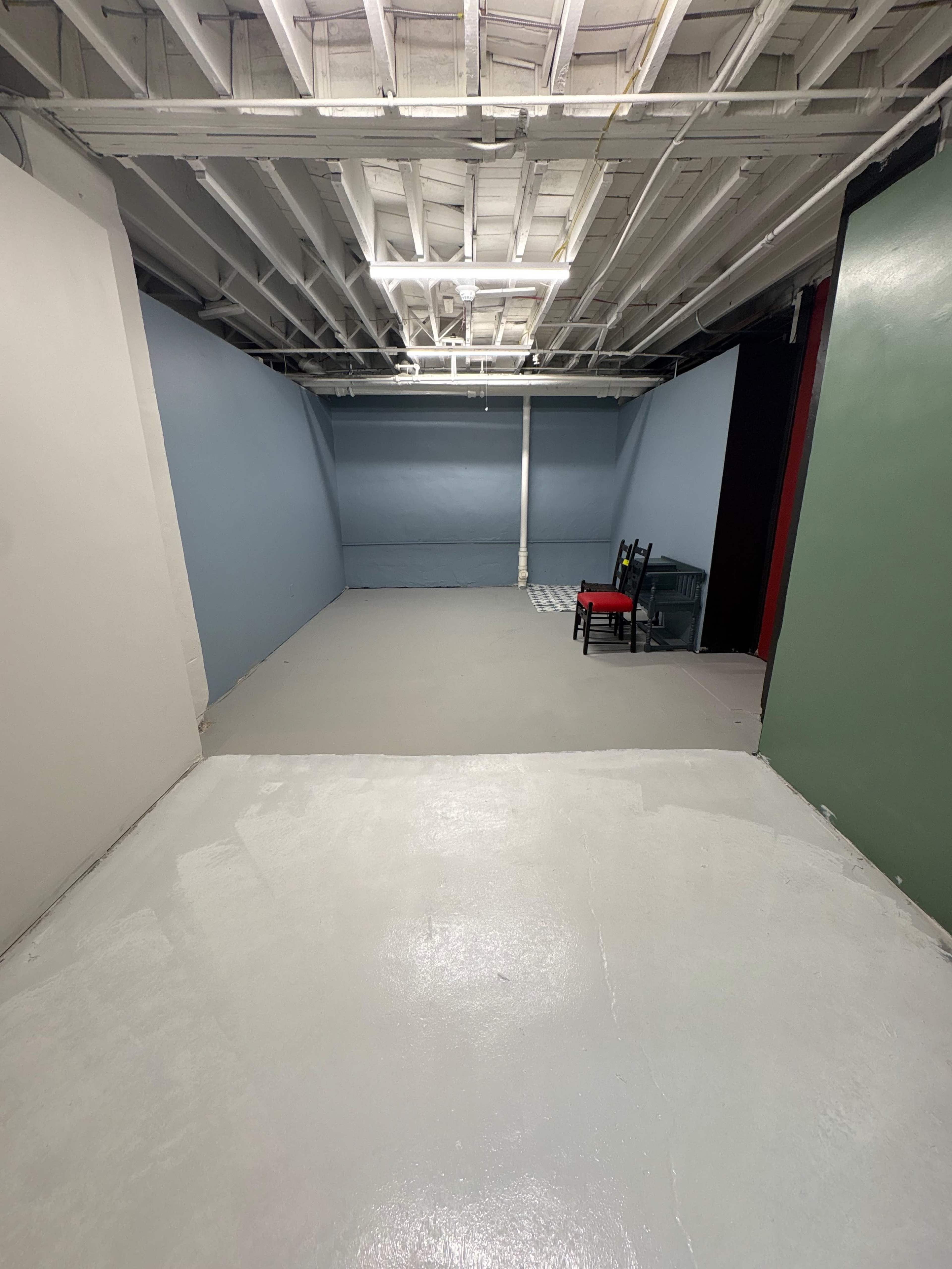 The image shows a spacious, unfinished basement with gray painted floors and blue walls, featuring a black chair and a small red table in one corner.