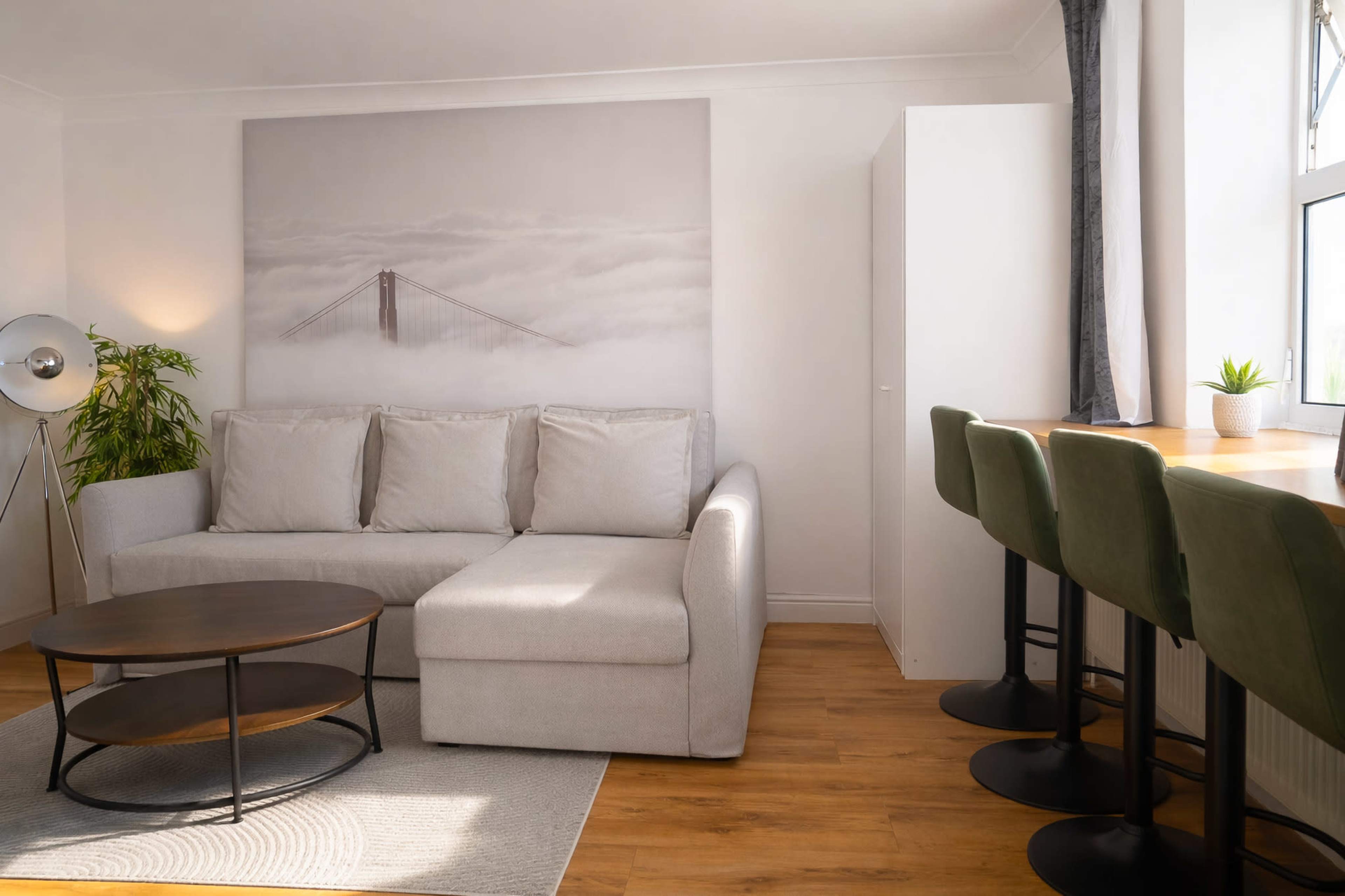 A light-colored living room features a sectional sofa, a round coffee table, a wall-mounted artwork of a bridge in fog, and a small dining area with bar stools.