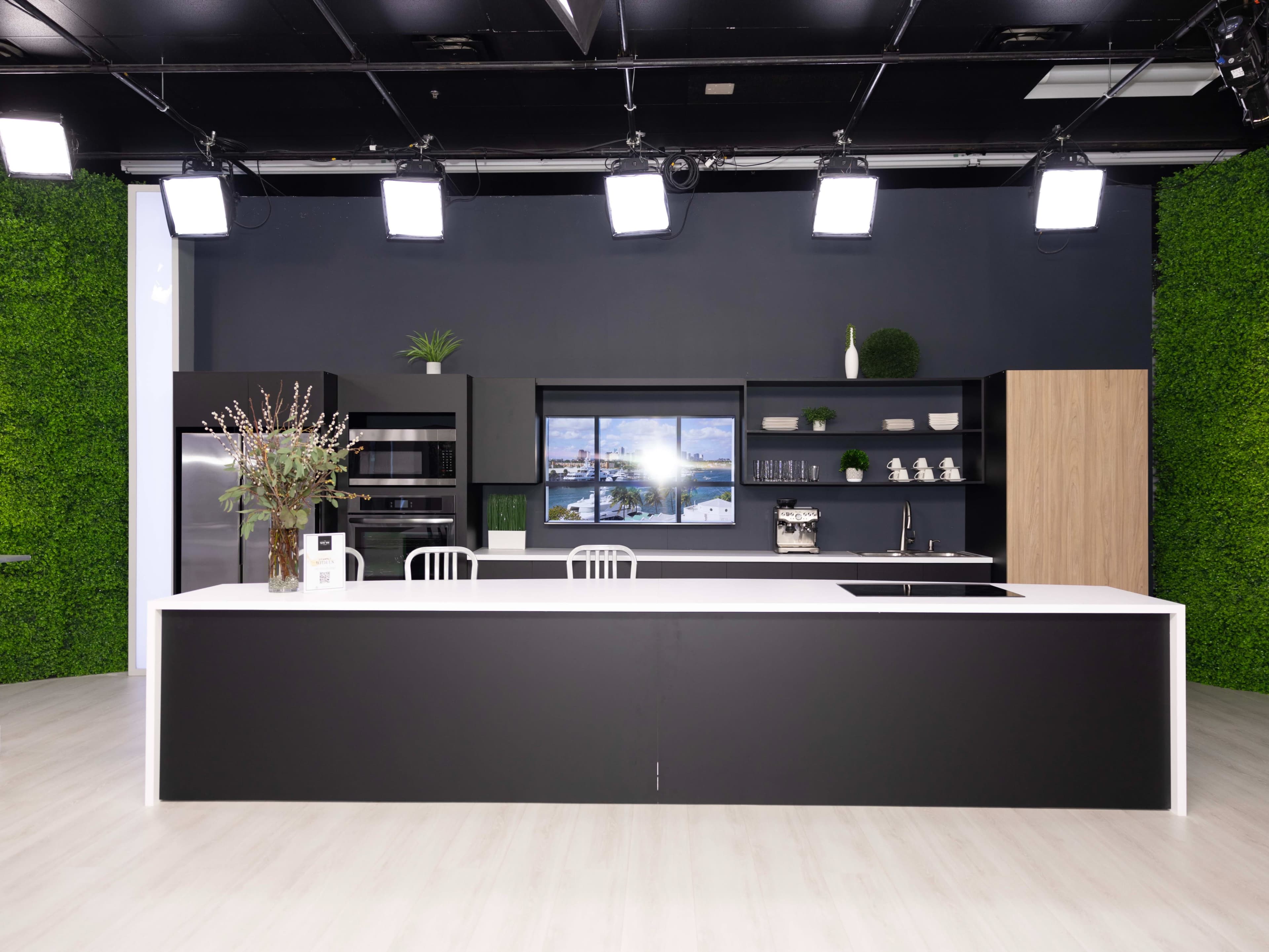 The image shows a modern kitchen set with a large island, stainless steel appliances, and a backdrop of green plants.