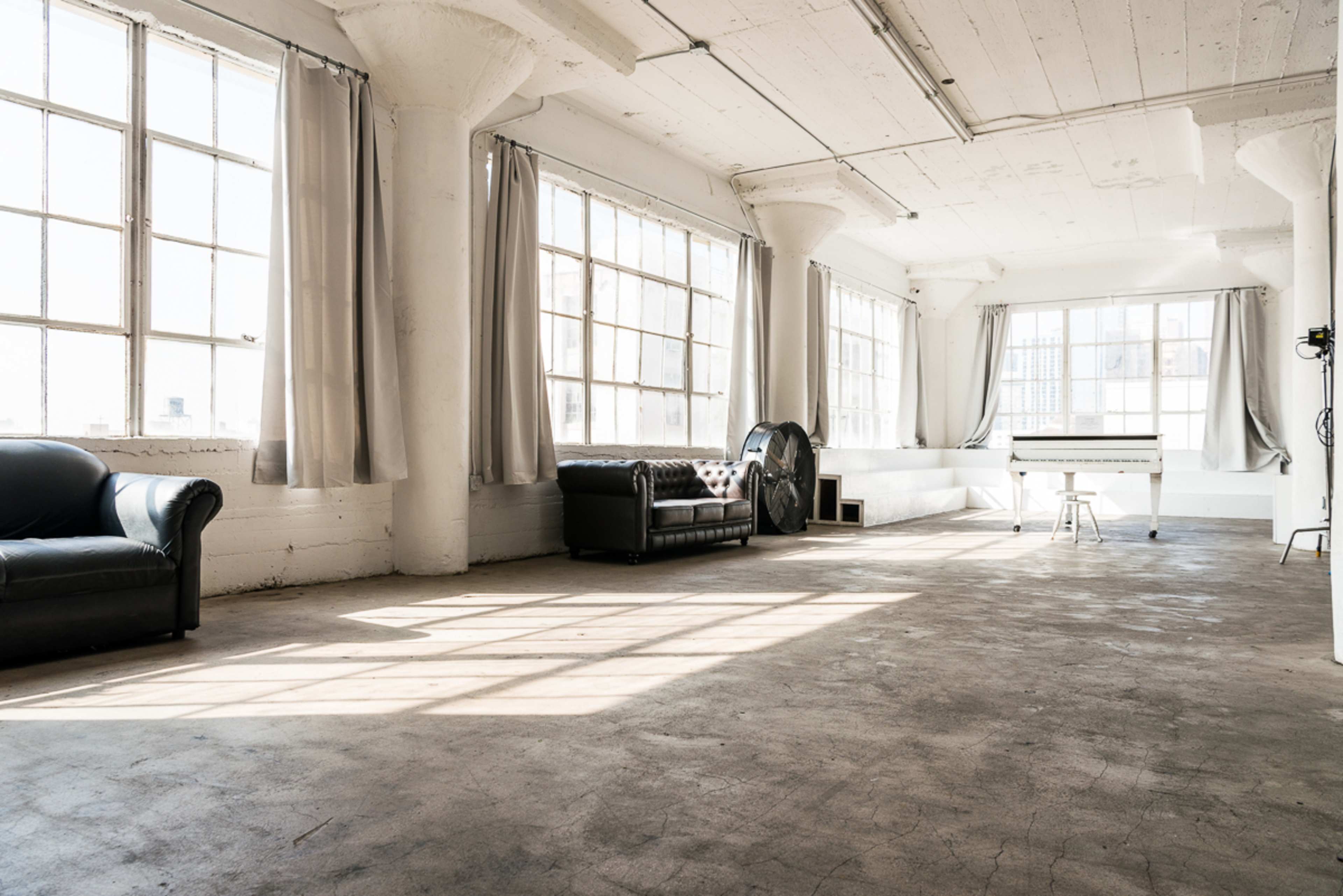The image shows a spacious, sunlit room with large windows, a grand piano, two leather couches, and bare concrete floors.