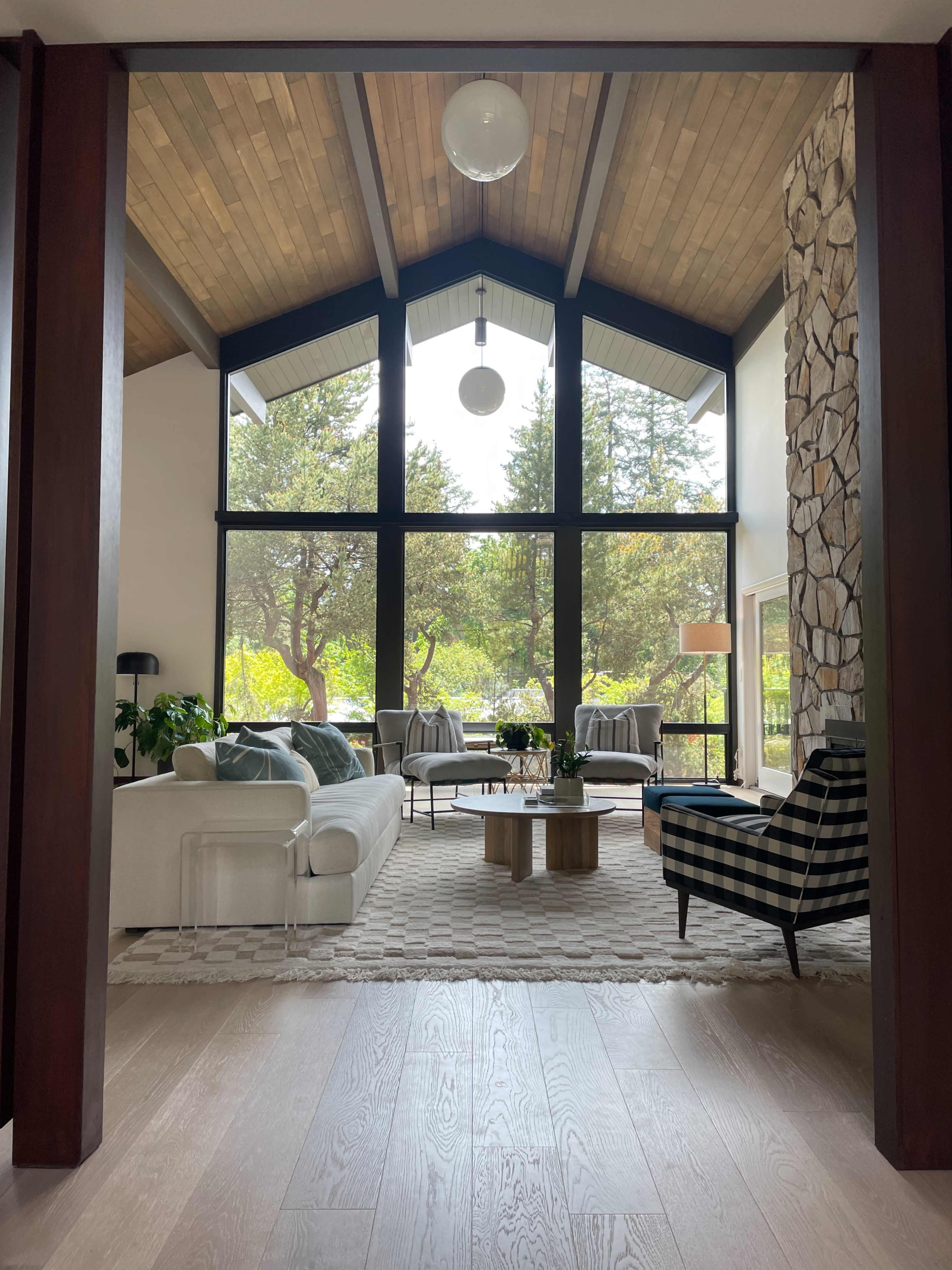 A modern living room with large windows overlooking a green landscape, featuring a light-colored sofa, a patterned armchair, and a coffee table on a textured rug.