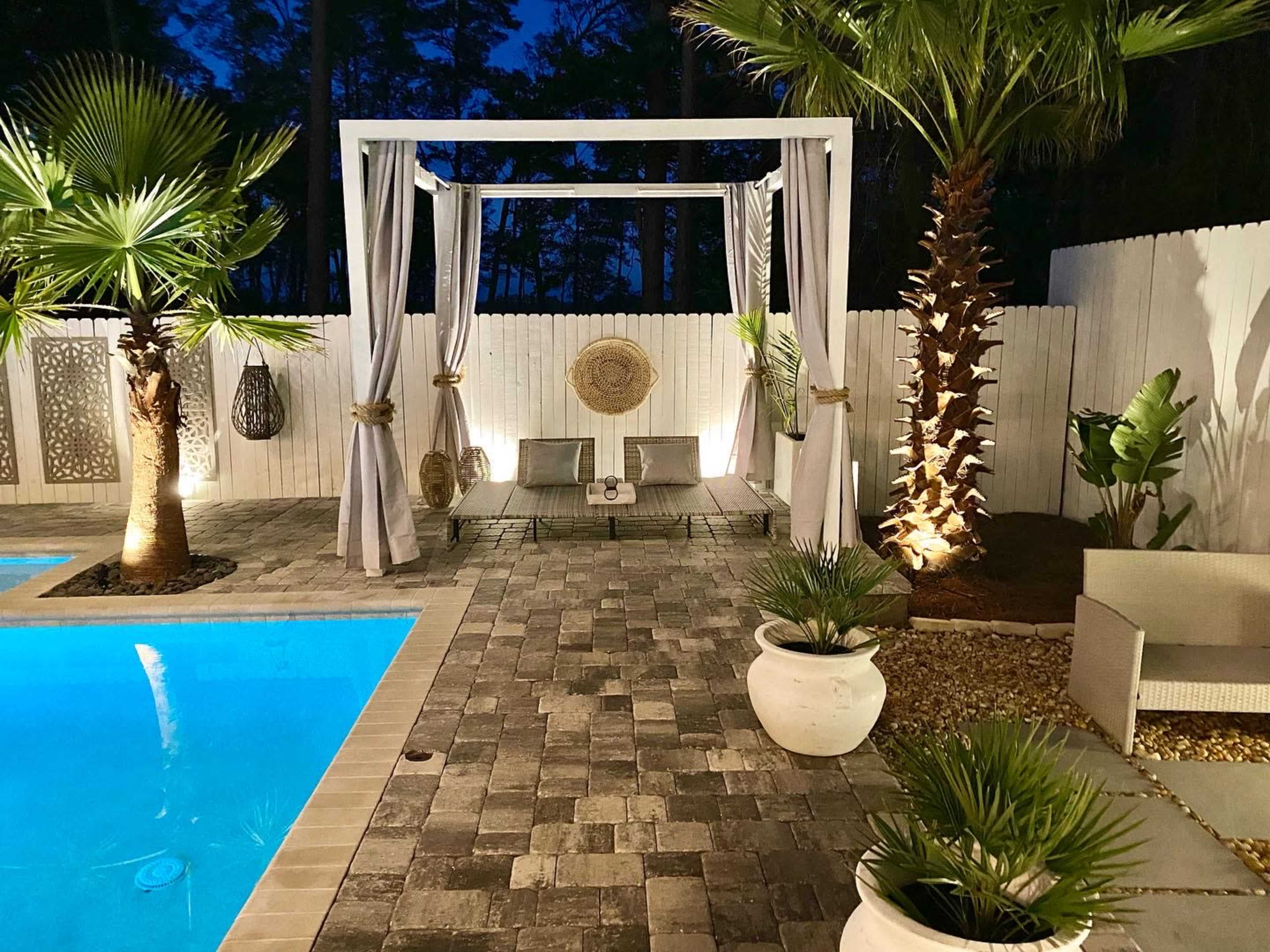 A backyard pool area at night, featuring a lit blue pool, a shaded seating structure with draped curtains, and landscaped greenery.