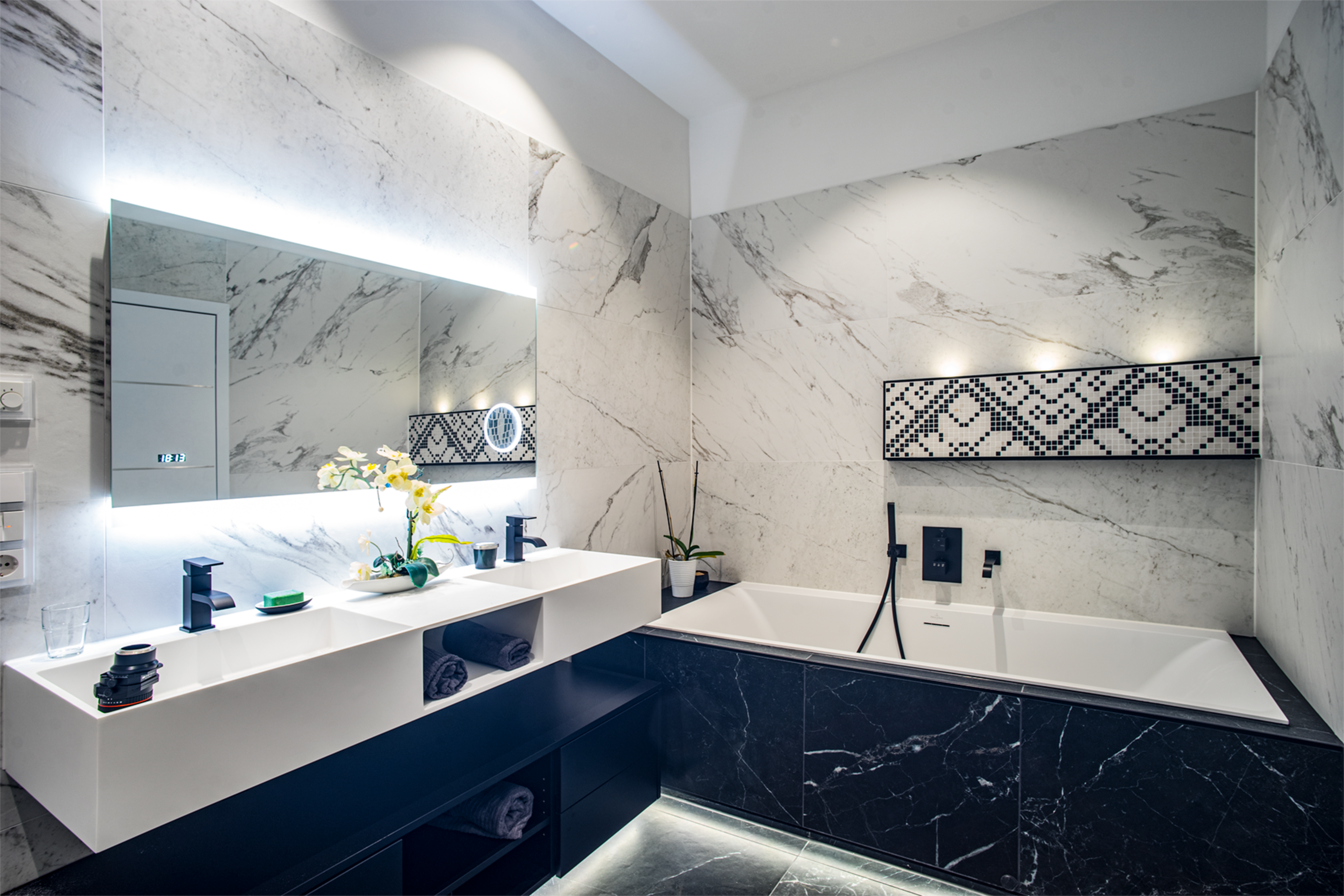 A modern bathroom features a marble-tiled wall, a double sink vanity with a large mirror, and a freestanding bathtub.