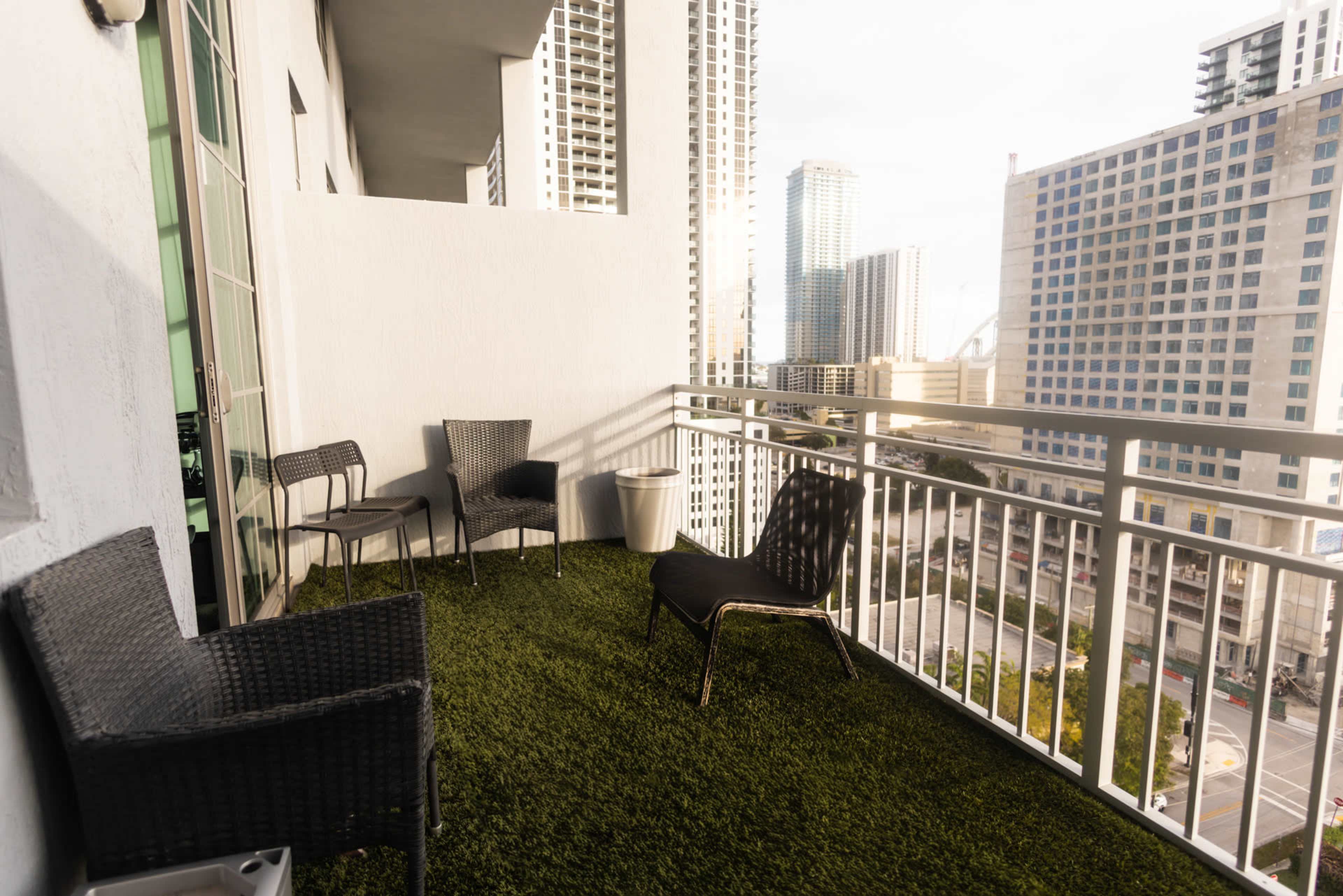 The image shows a balcony with artificial grass, featuring four chairs and a small table, overlooking a city skyline.