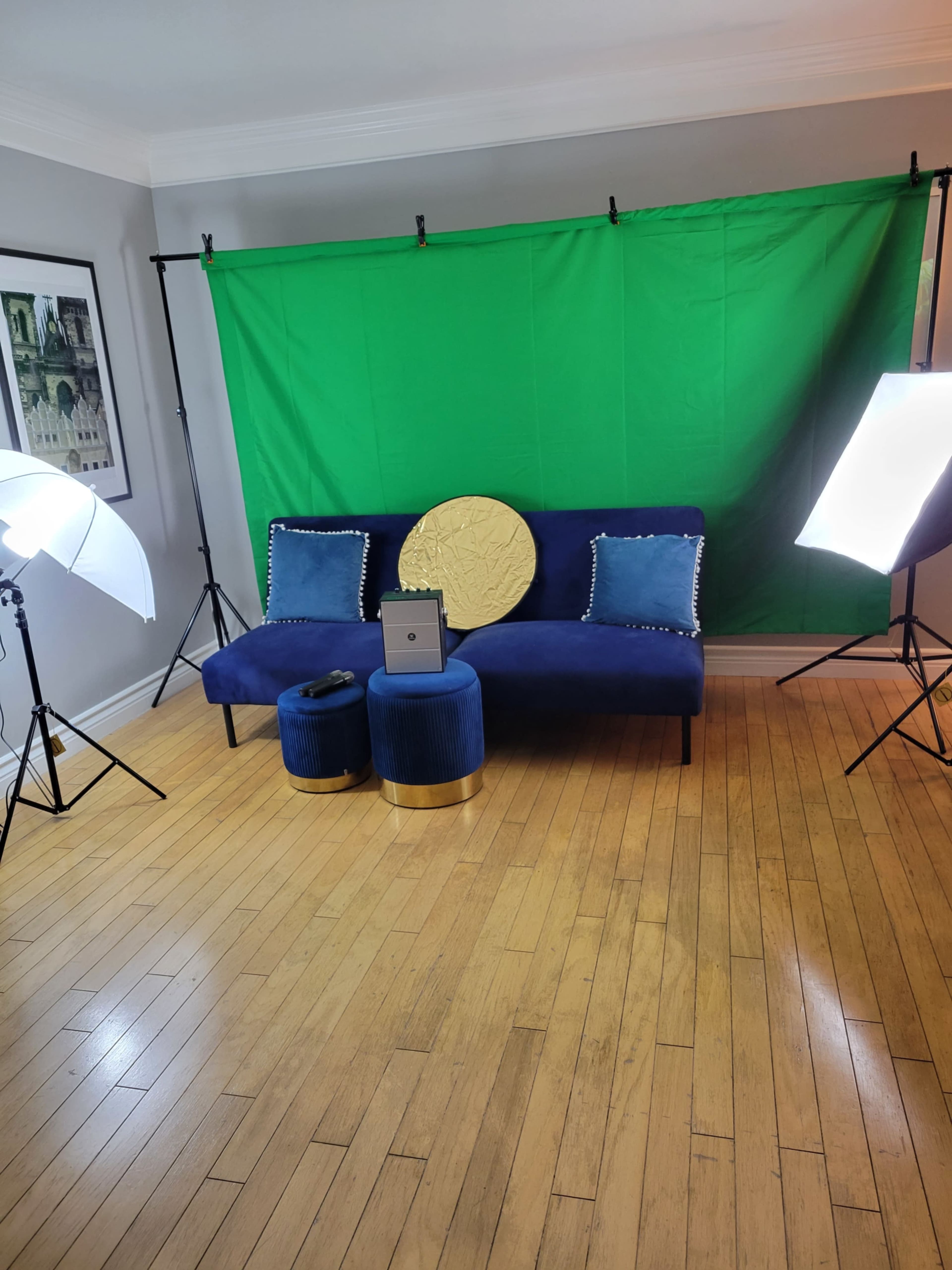 The image shows a blue couch with decorative pillows, two blue ottomans, a gold circular pillow, and a green backdrop, all set up with two bright lights on tripods in a room with wooden flooring.