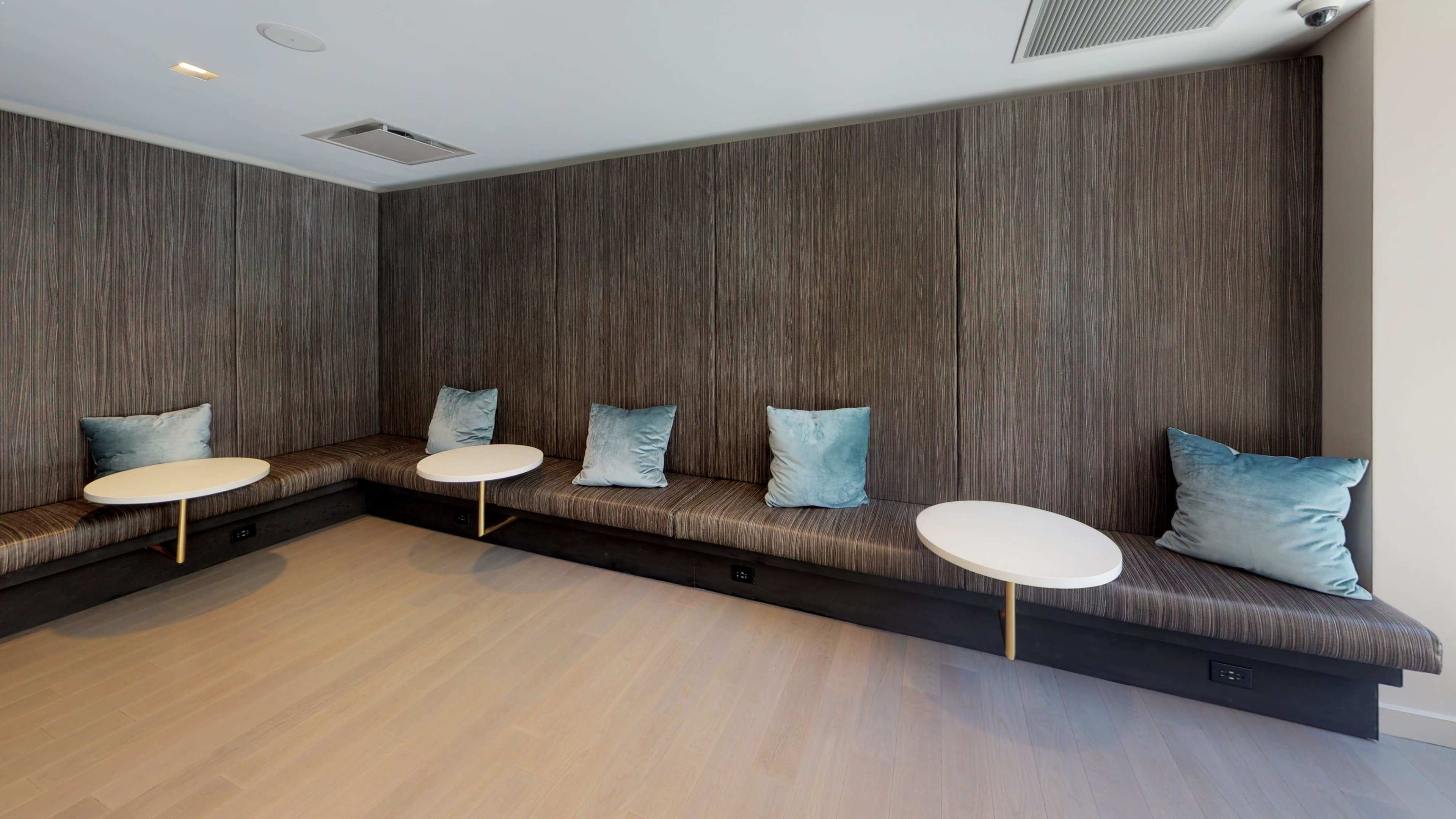 A modern, minimalist seating area with a bench, several blue cushions, and small round tables against a dark wooden wall.