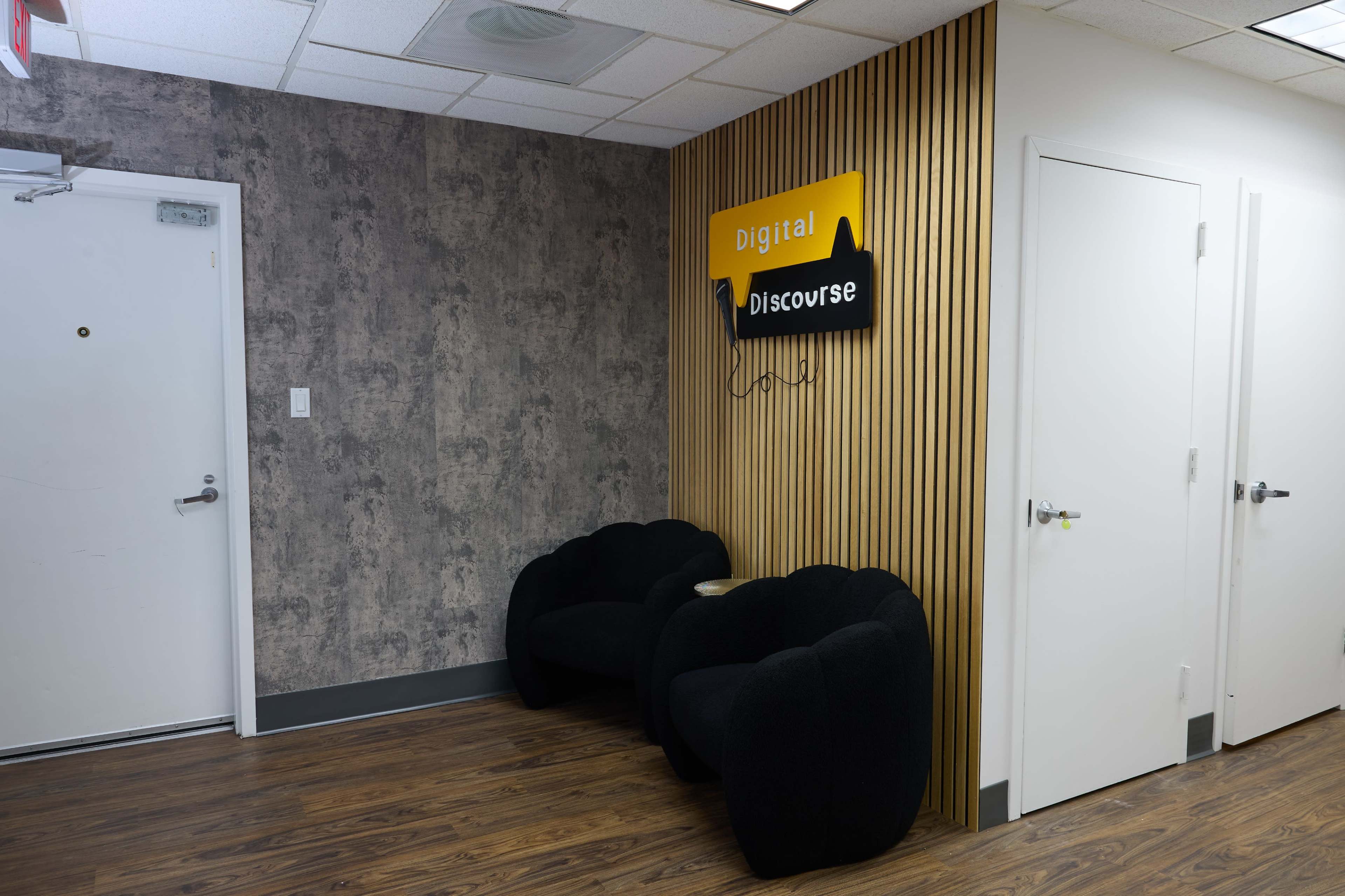 The image shows a small waiting area with two black lounge chairs against a textured wall, featuring a sign that reads "Digital Discourse" above.