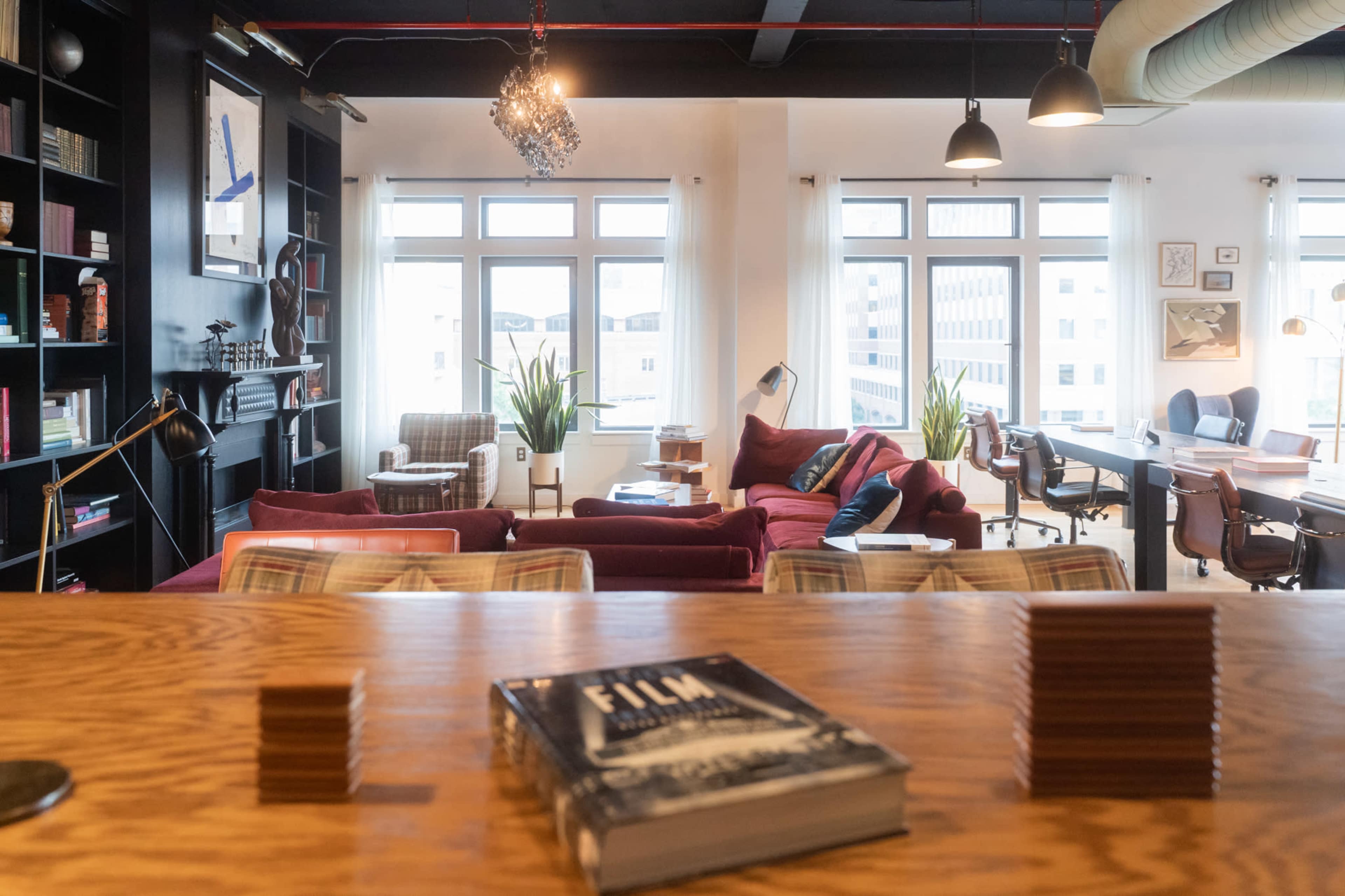 The image shows a cozy, modern office space with a mix of seating areas, bookshelves, and large windows allowing natural light.