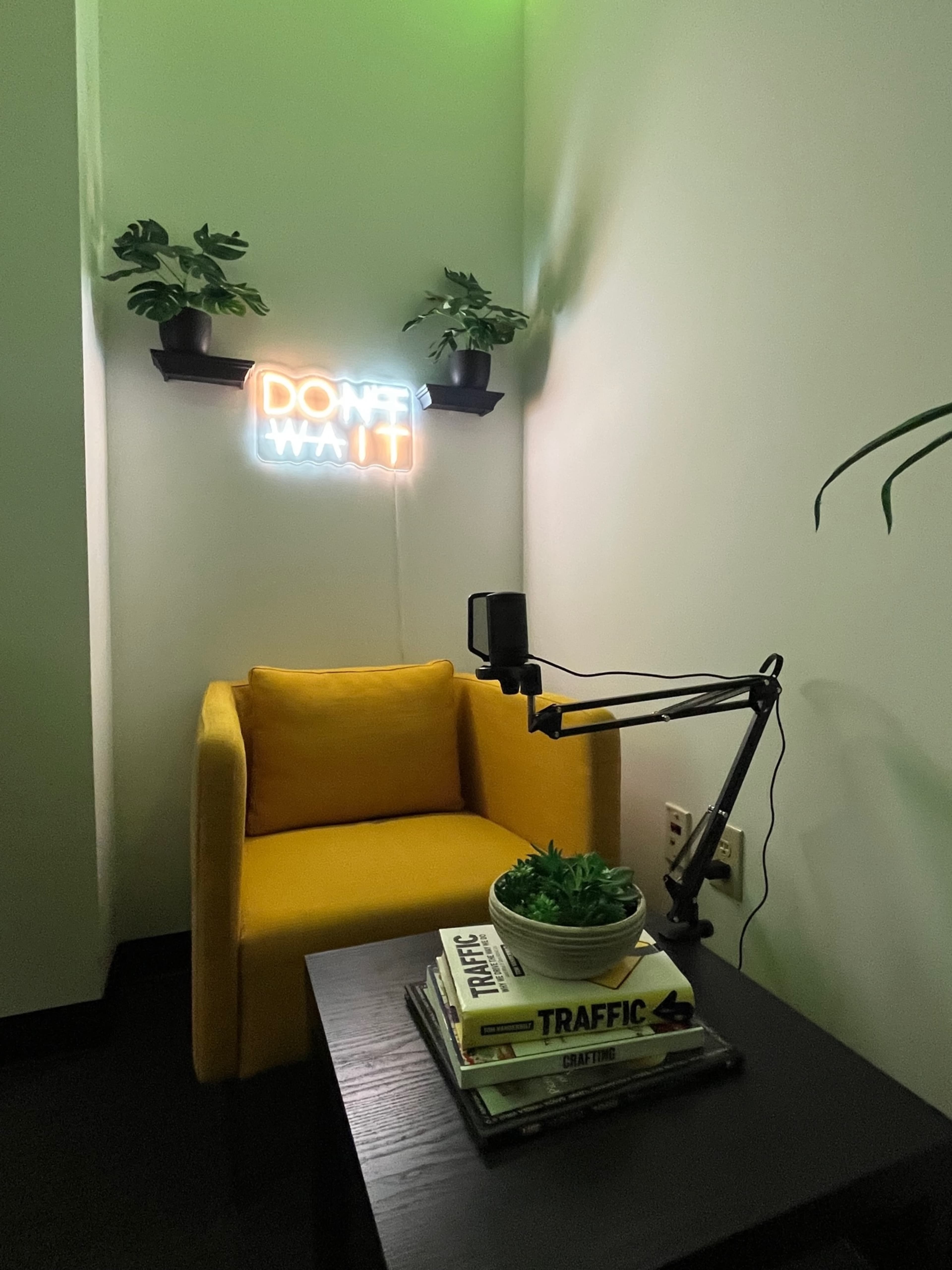 A yellow armchair next to a black table with stacked books and a potted plant, set against a wall featuring a neon sign that reads "DON'T WANT IT."
