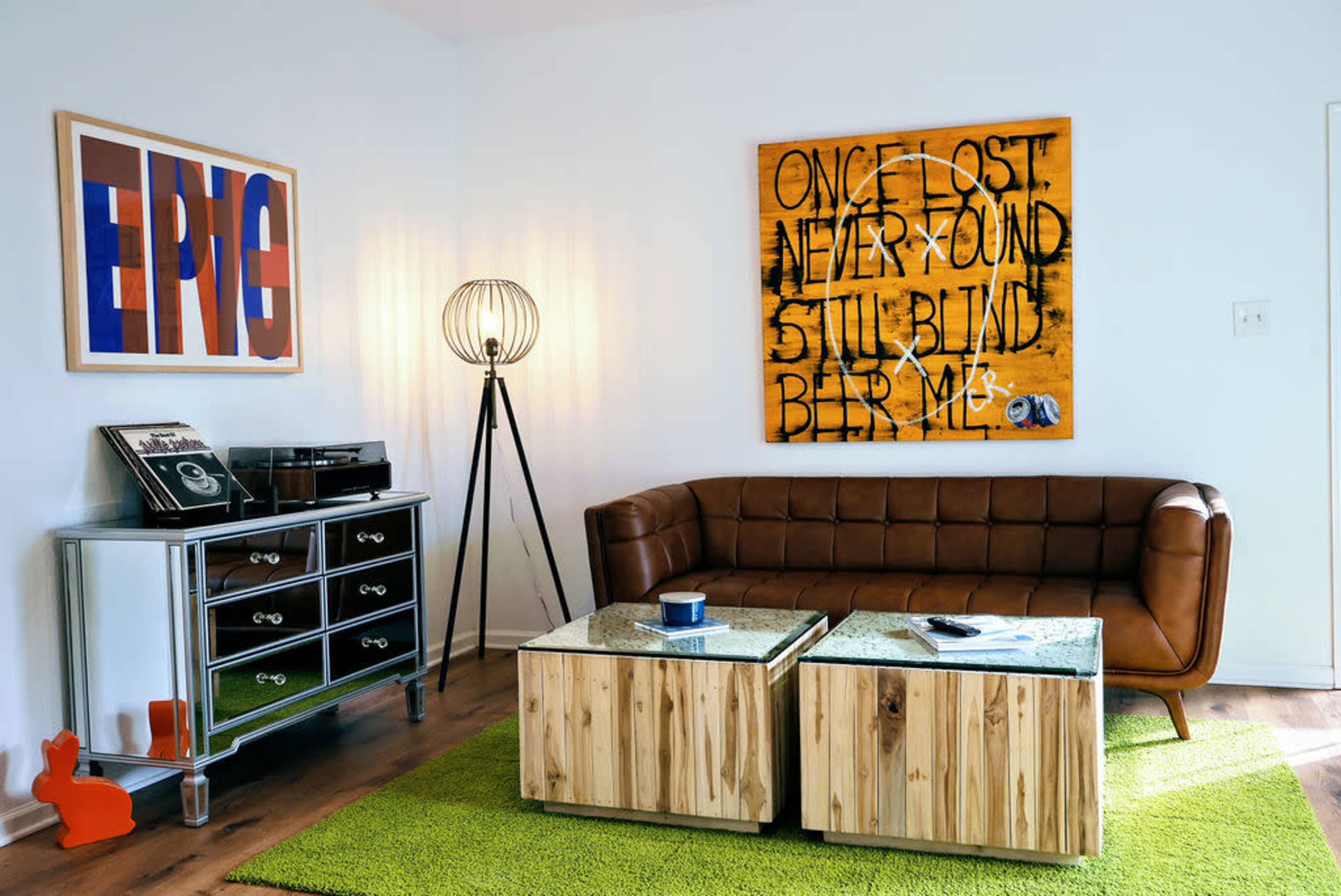 The image features a modern living room with a brown leather sofa, a wooden coffee table, a silver side table with a record player, and a large art piece on the wall.