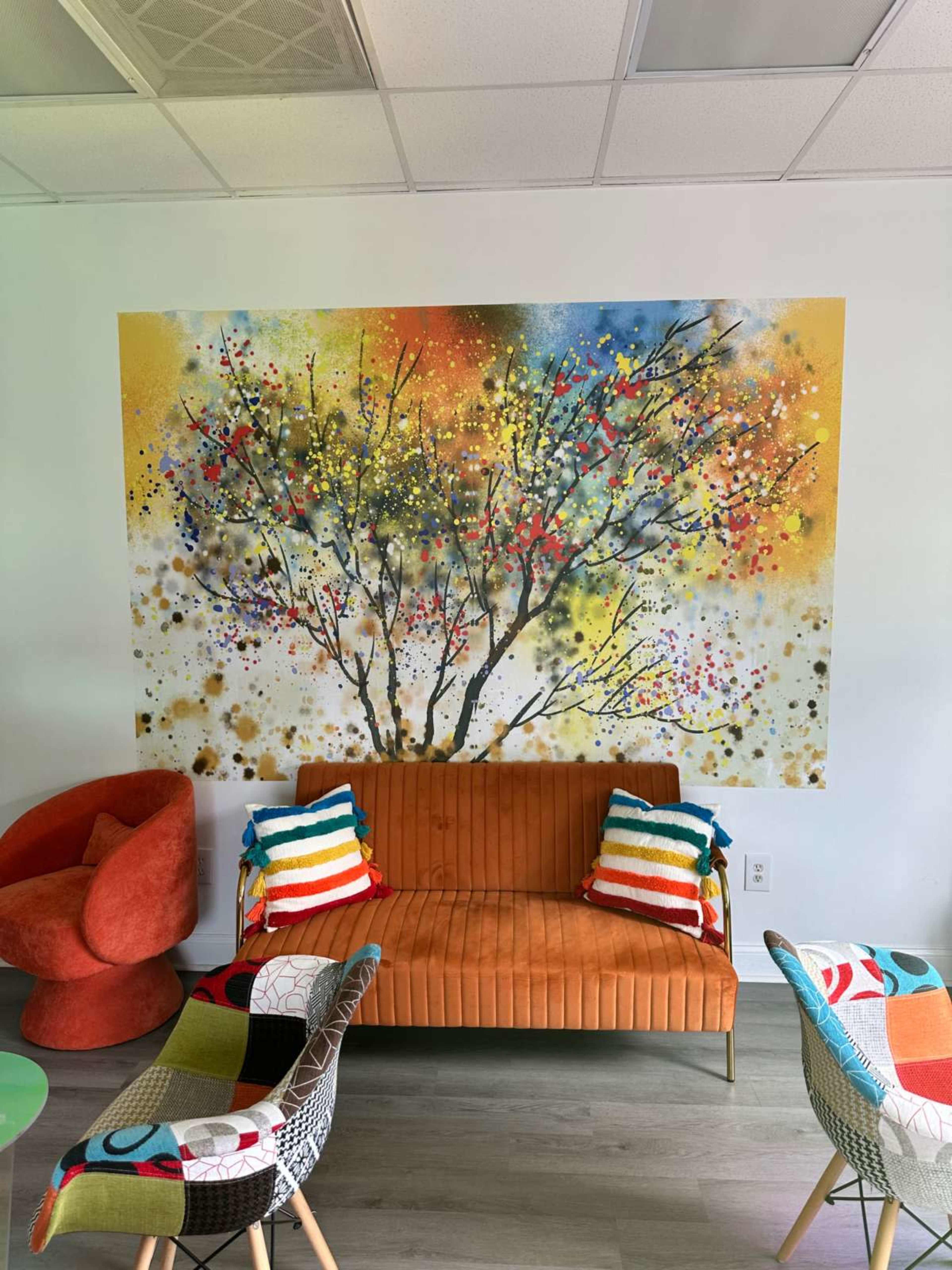 The image showcases a colorful mural of a tree on a wall behind an orange couch adorned with striped cushions, surrounded by uniquely patterned chairs and a glass table.