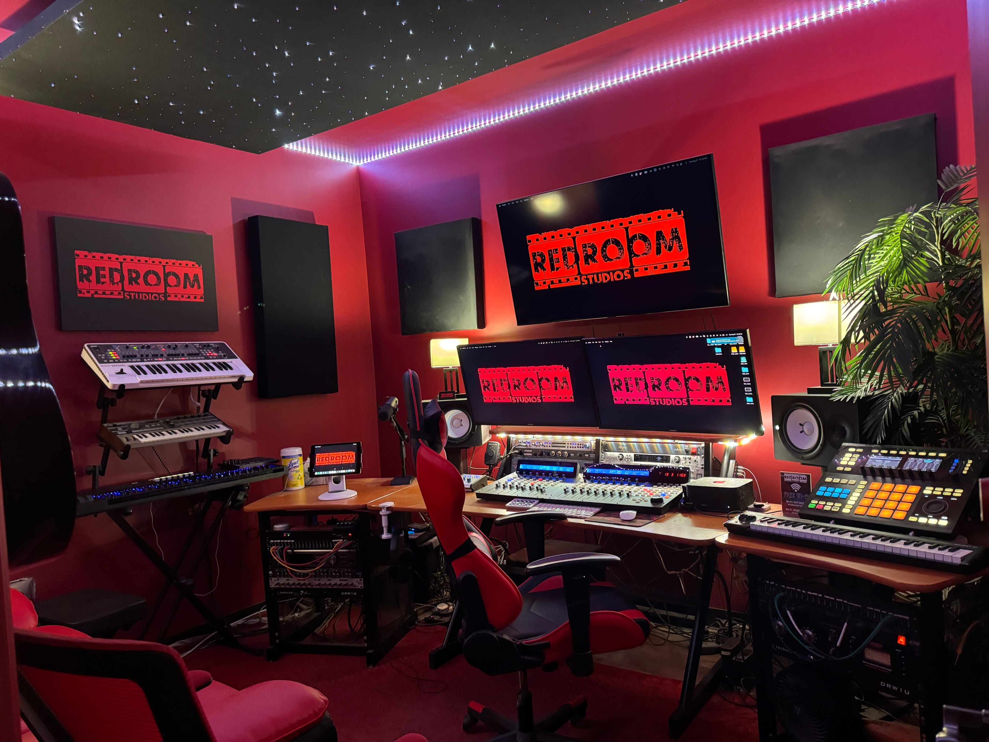 A music production studio with various keyboards, sound equipment, and monitors arranged around a central workstation, all set in a red-themed room with starry ceiling lights.