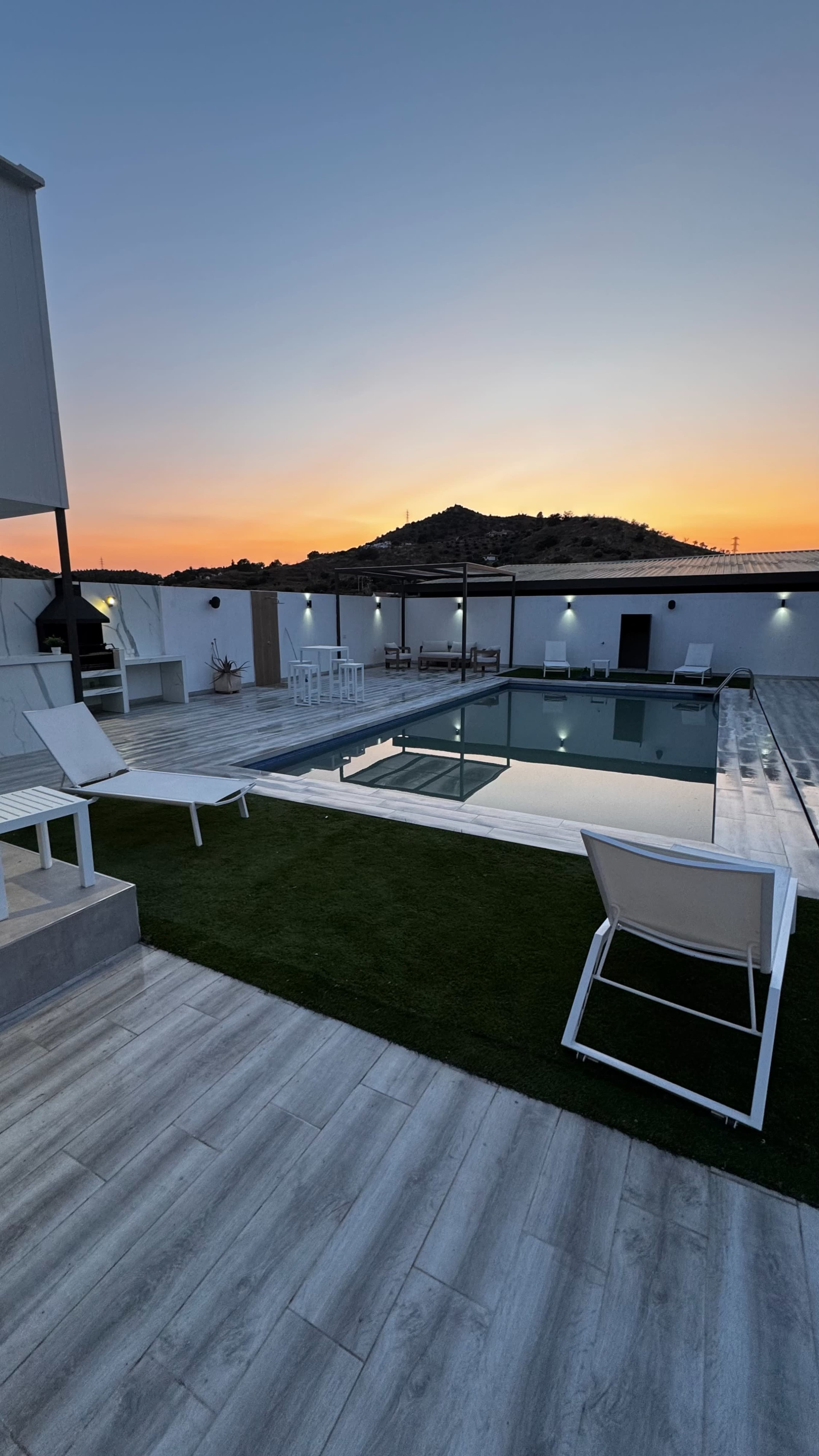 The image shows a pool area with white lounge chairs, a landscaped lawn, and a clear view of a sunset over a distant hill.