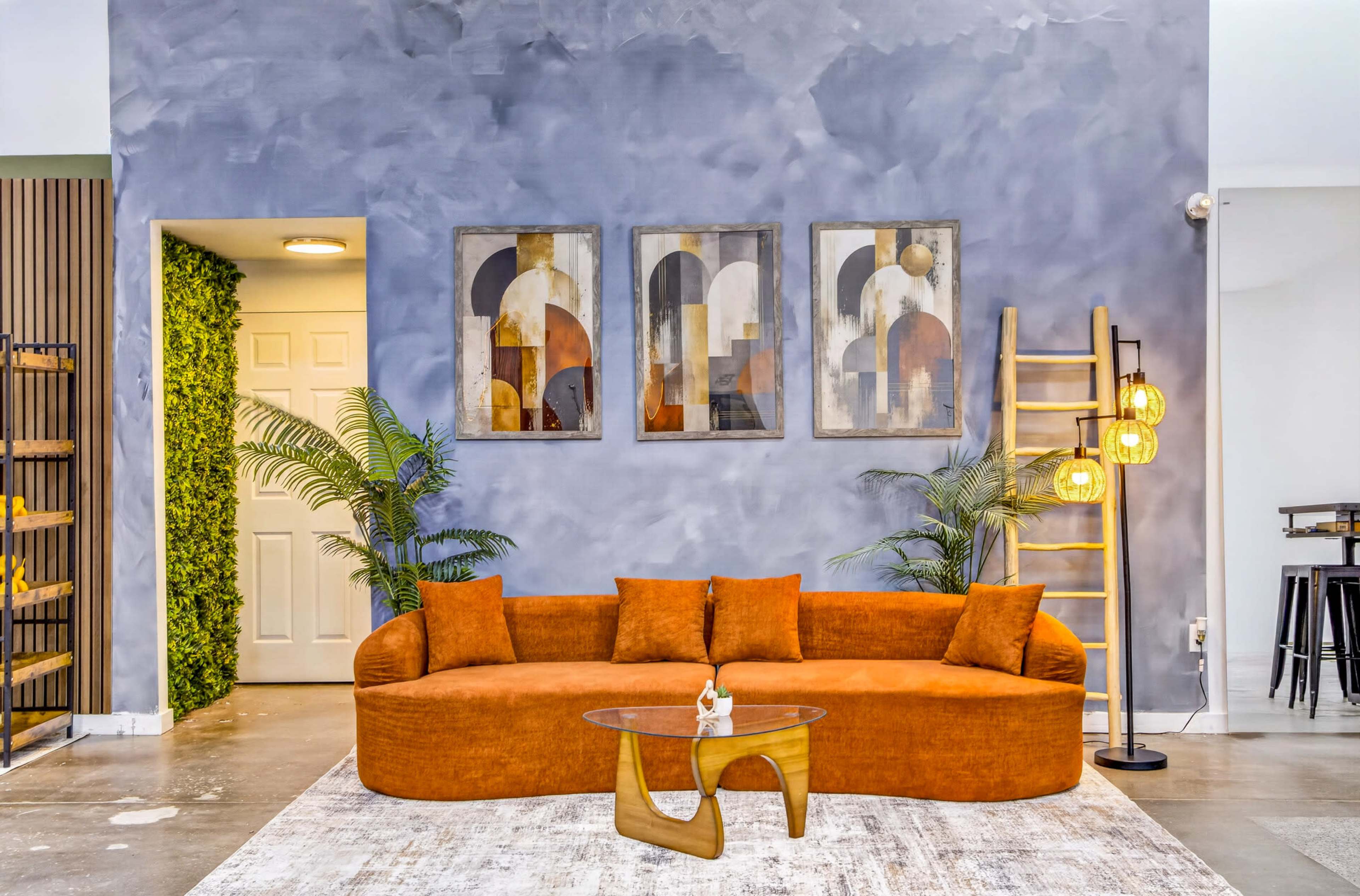 The image shows a modern living room featuring an orange curved couch, a glass coffee table, and abstract artwork on the wall, with indoor plants and decorative lighting.