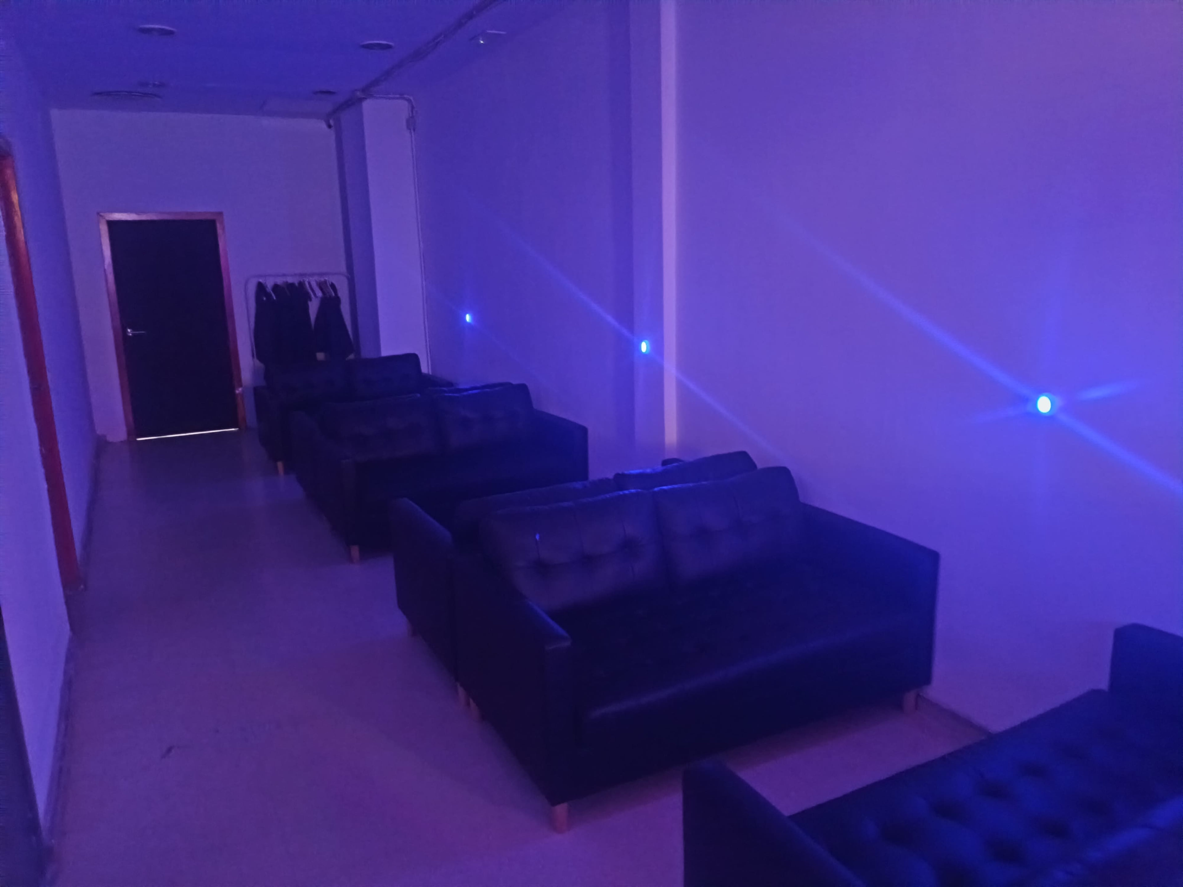 The image shows a dimly lit lounge area with several dark sofas arranged in rows along a wall, with blue lights illuminating the space.