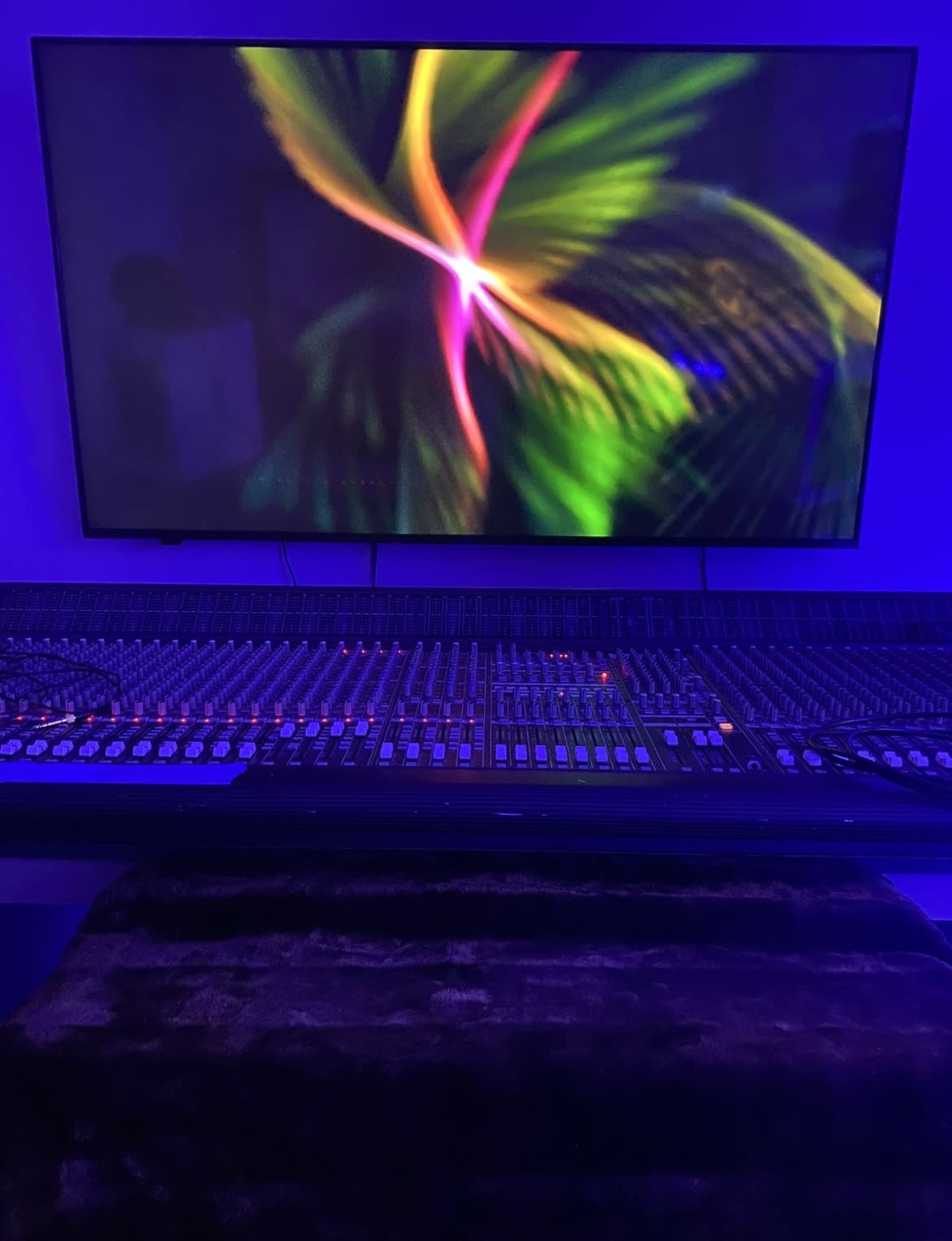 A digital audio mixing console is positioned in front of a large wall-mounted monitor displaying colorful abstract graphics.