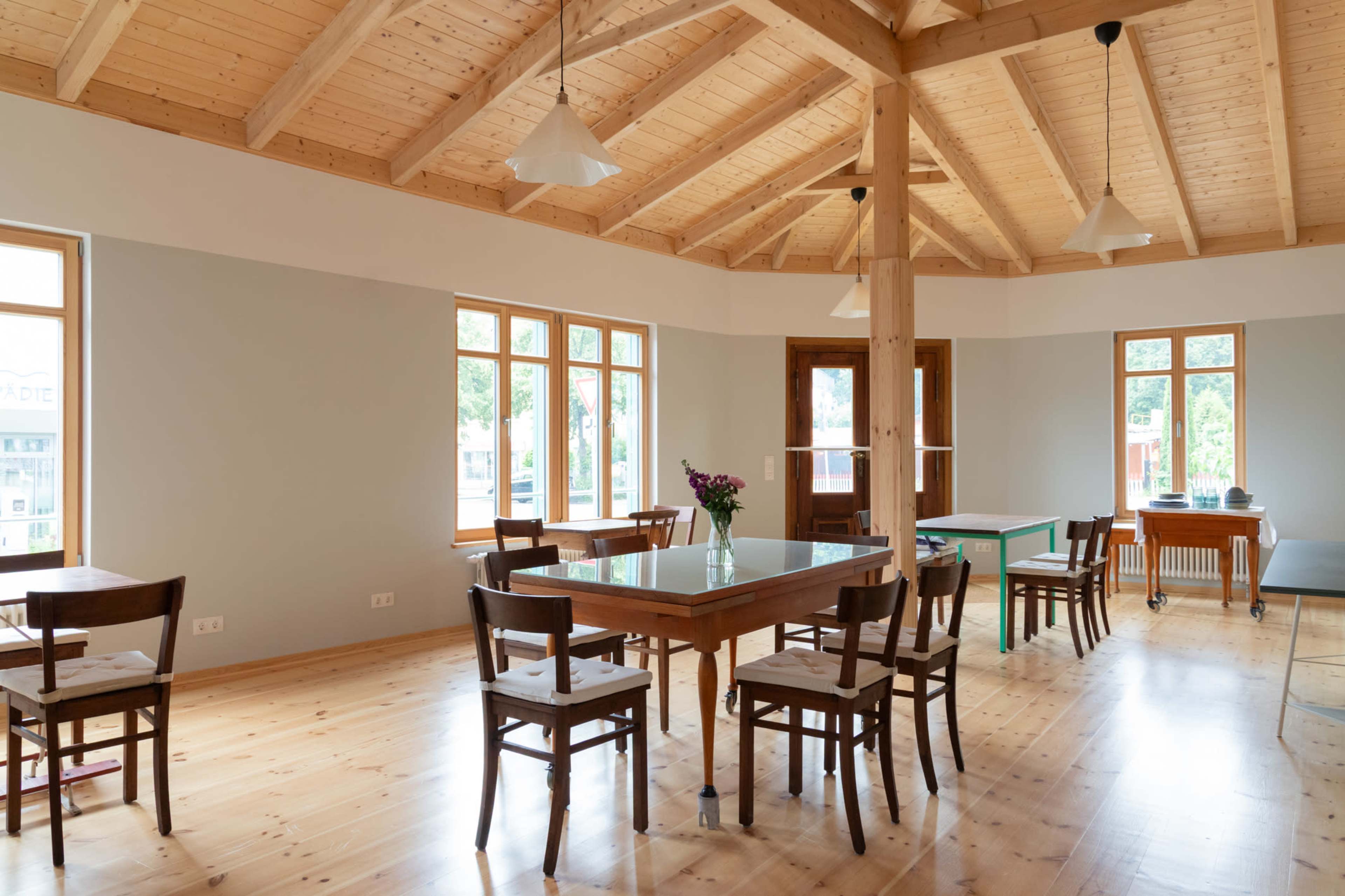 The image shows a spacious, well-lit room with wooden tables and chairs arranged symmetrically around large windows and a wooden ceiling.