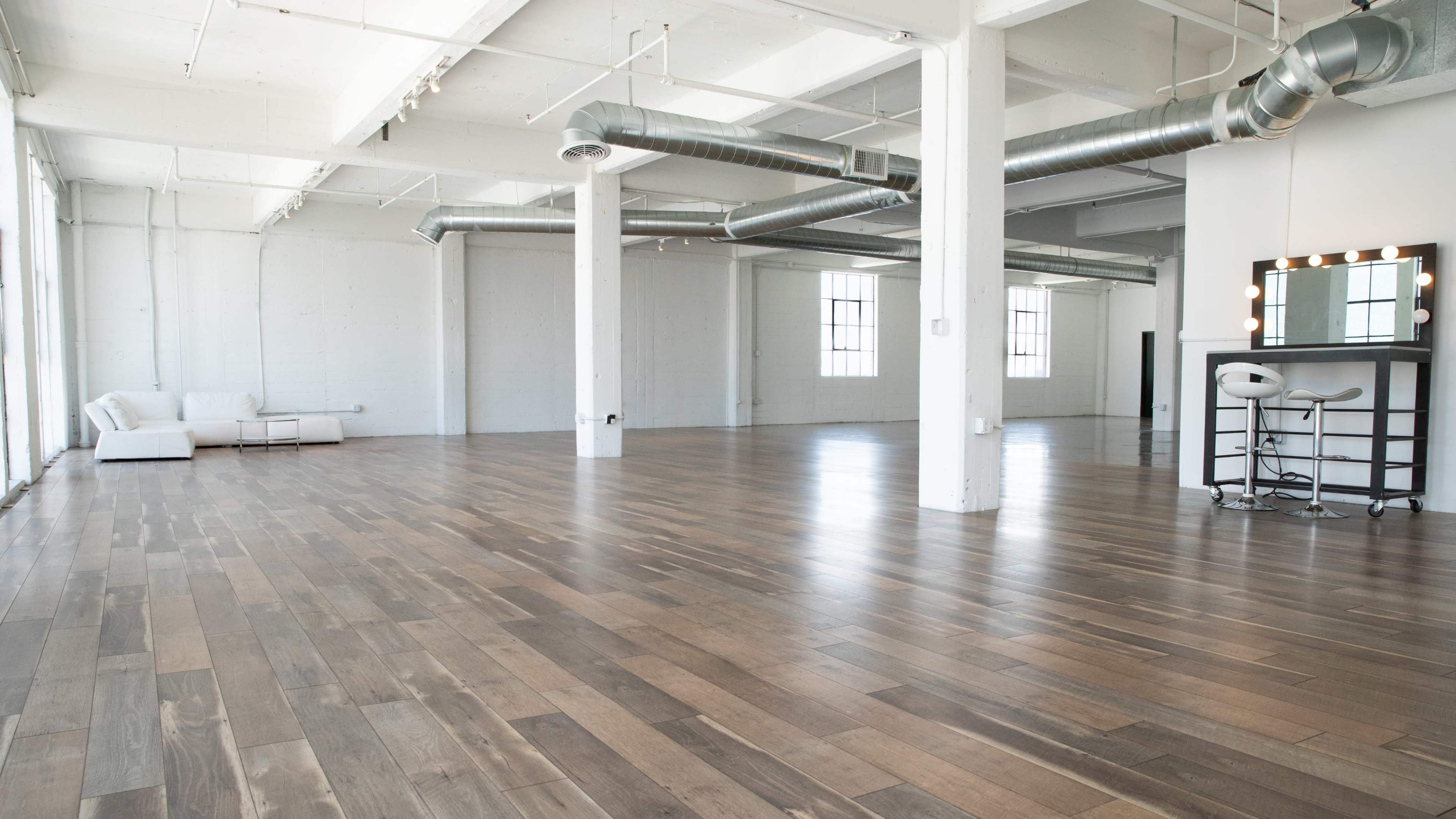 The image shows a spacious, empty loft with hardwood flooring and large windows, featuring a white sofa and a vanity area with a mirror and stool.