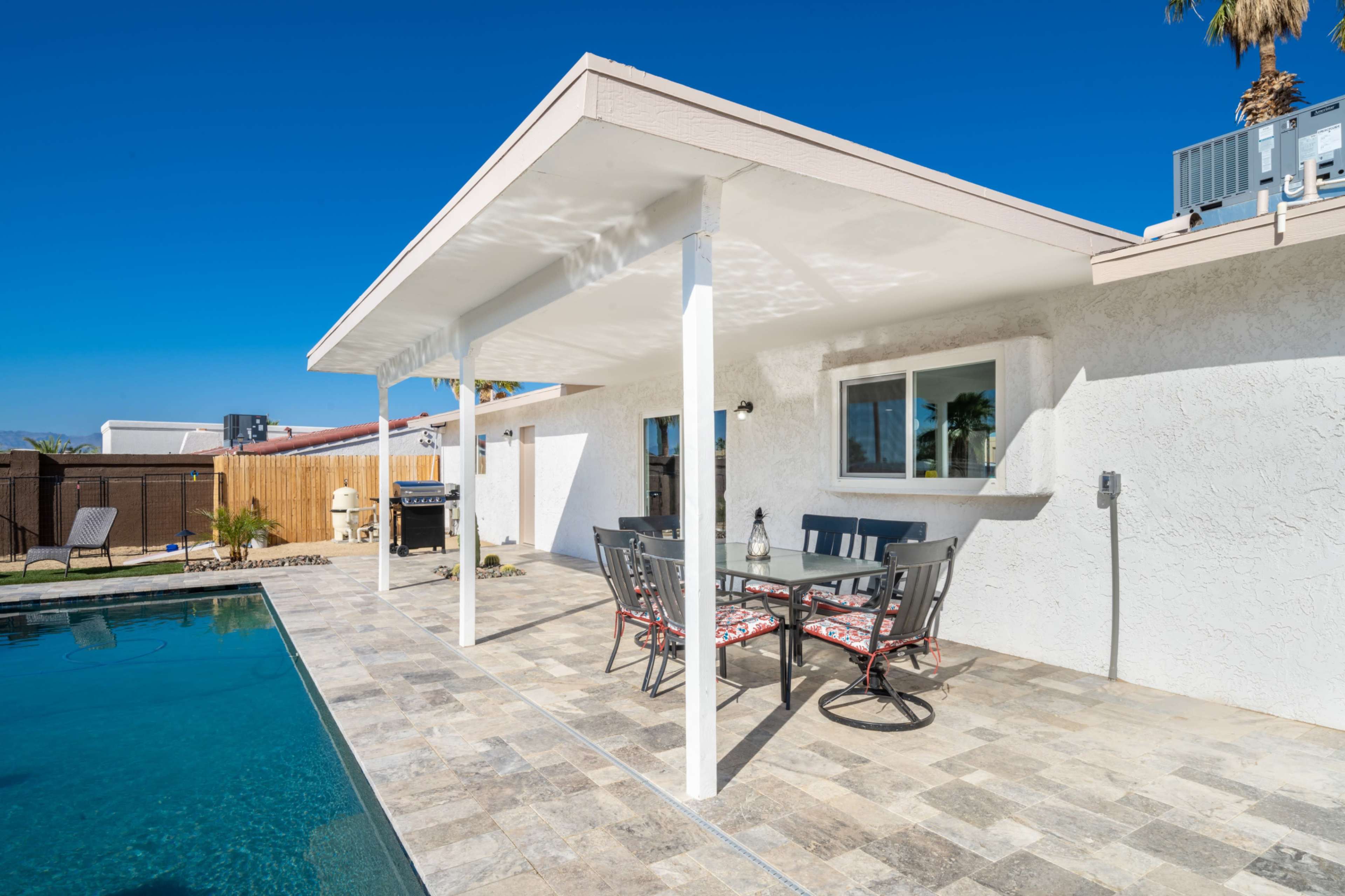 A patio with a dining table and chairs overlooks a swimming pool surrounded by stone tiles.