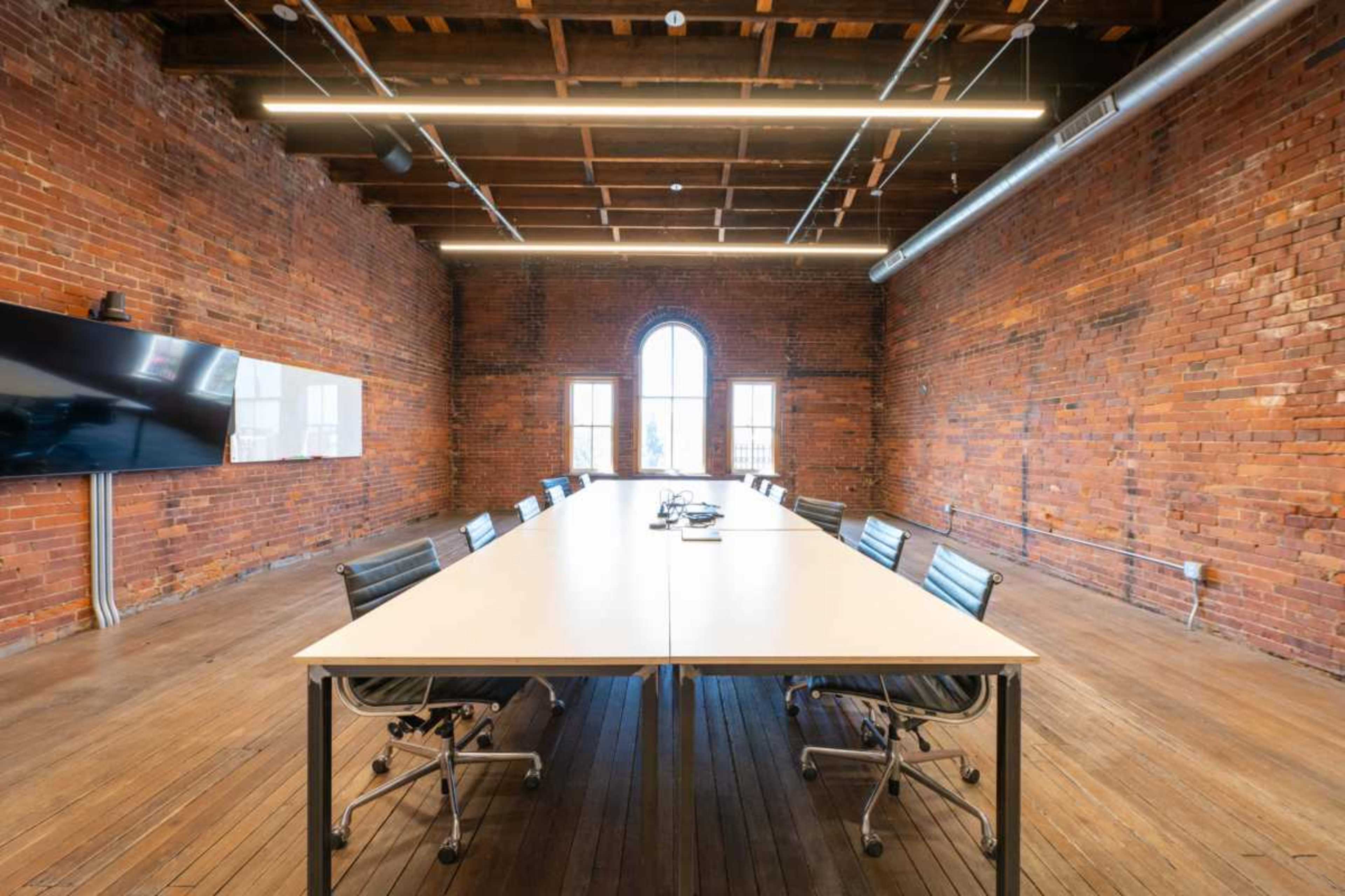 A long table with chairs is set up in a spacious conference room that features exposed brick walls and a large window.