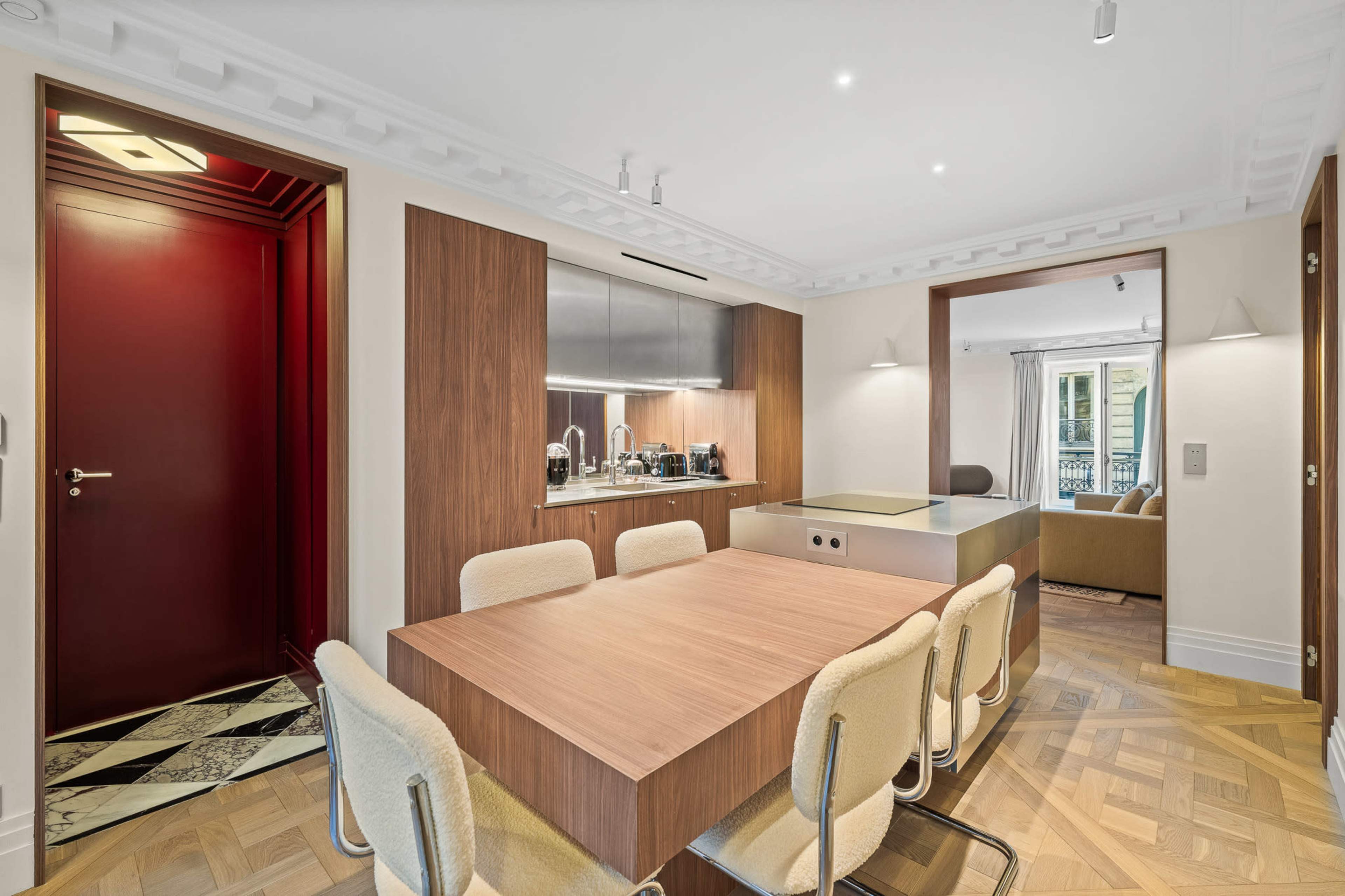 The image shows a modern kitchen and dining area featuring a wooden dining table surrounded by beige chairs, with a sleek kitchen setup in the background and a red door leading to another room.