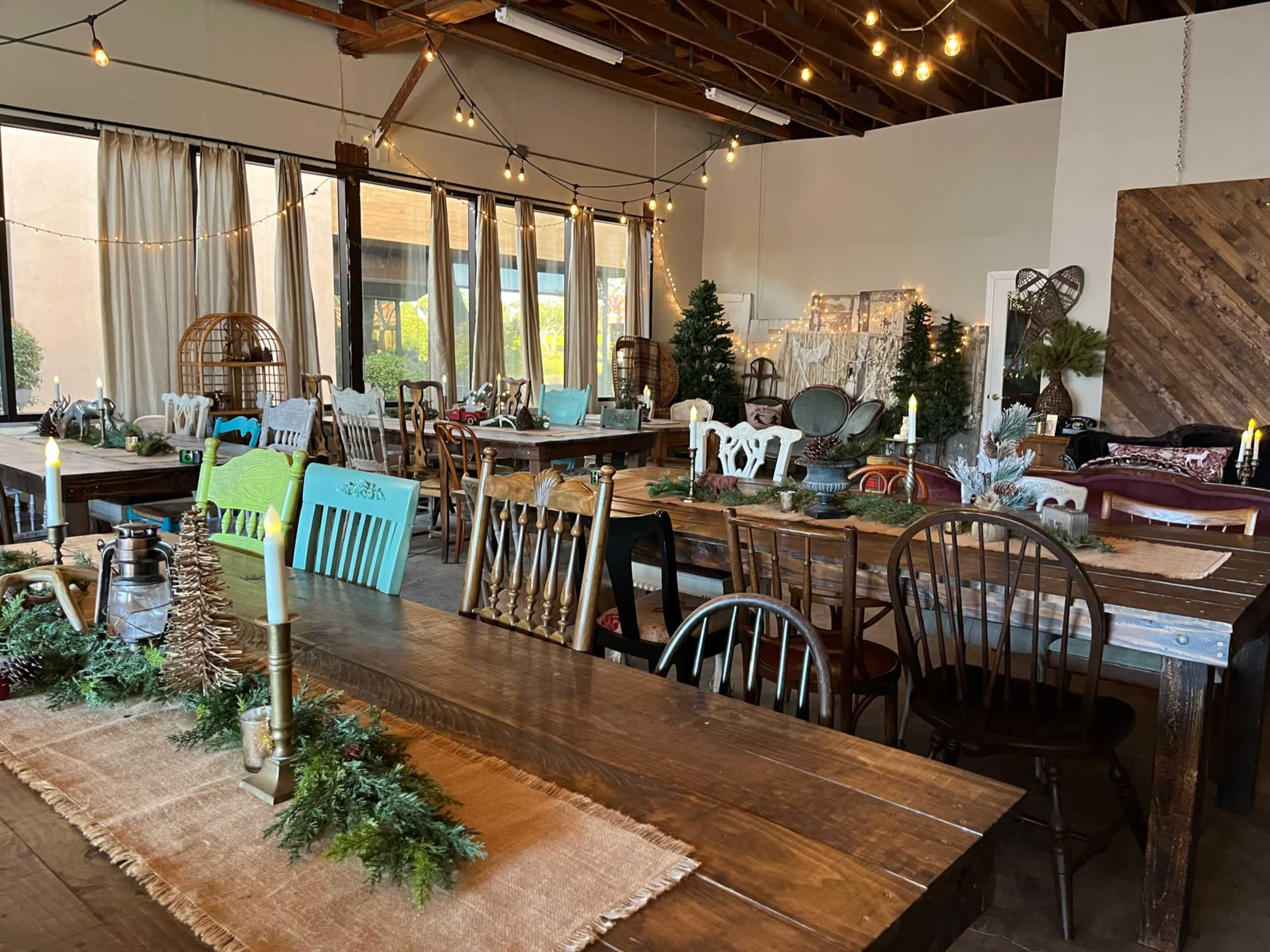 The image shows a spacious, decorated event room filled with various wooden tables and chairs, adorned with greenery and soft lighting.