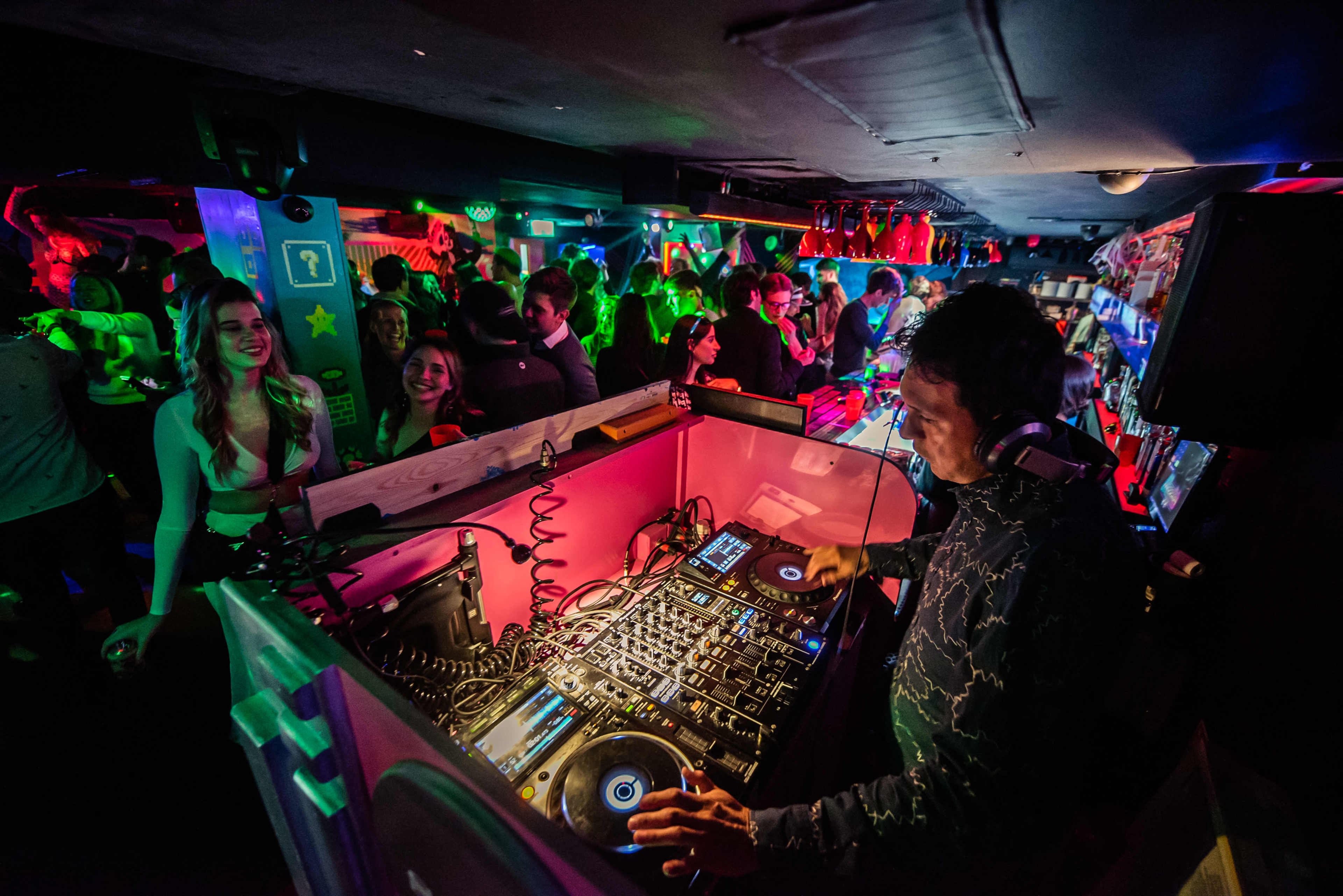 A DJ plays music at a lively nightclub filled with colorful lights and people dancing.