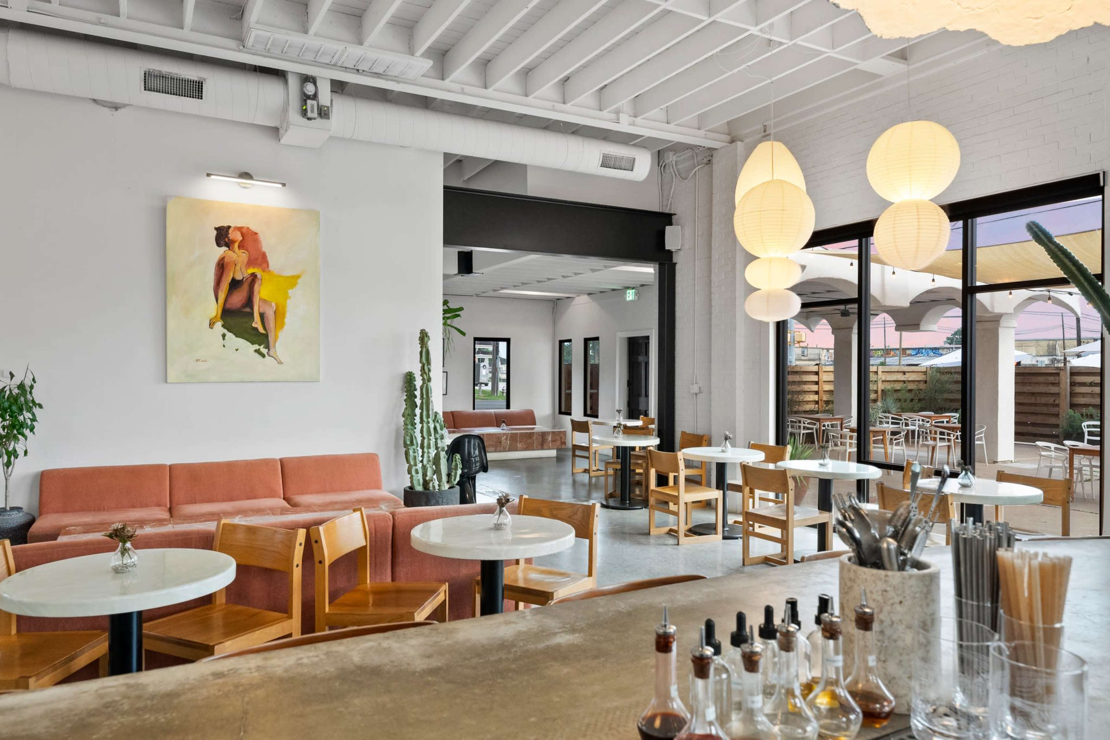 The image shows a modern cafe with a light-colored interior, featuring a mix of wooden furniture, a large artwork on the wall, and illuminated paper lanterns.