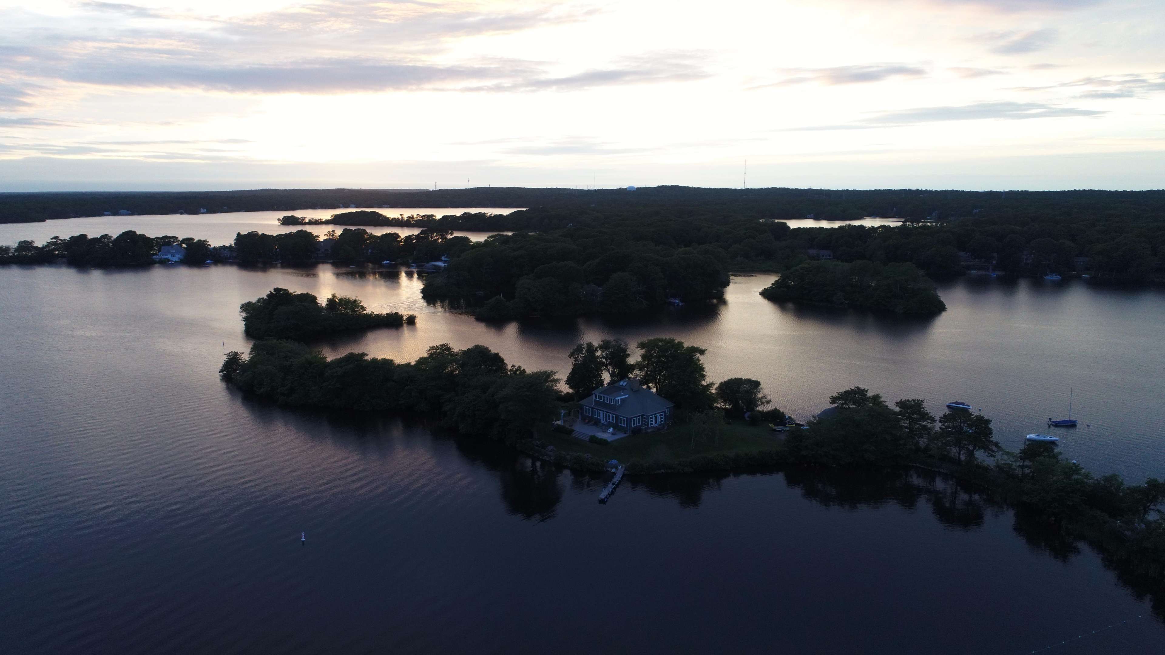 An aerial view shows a tranquil lake surrounded by scattered islands and a house situated on one of the islands at sunset.