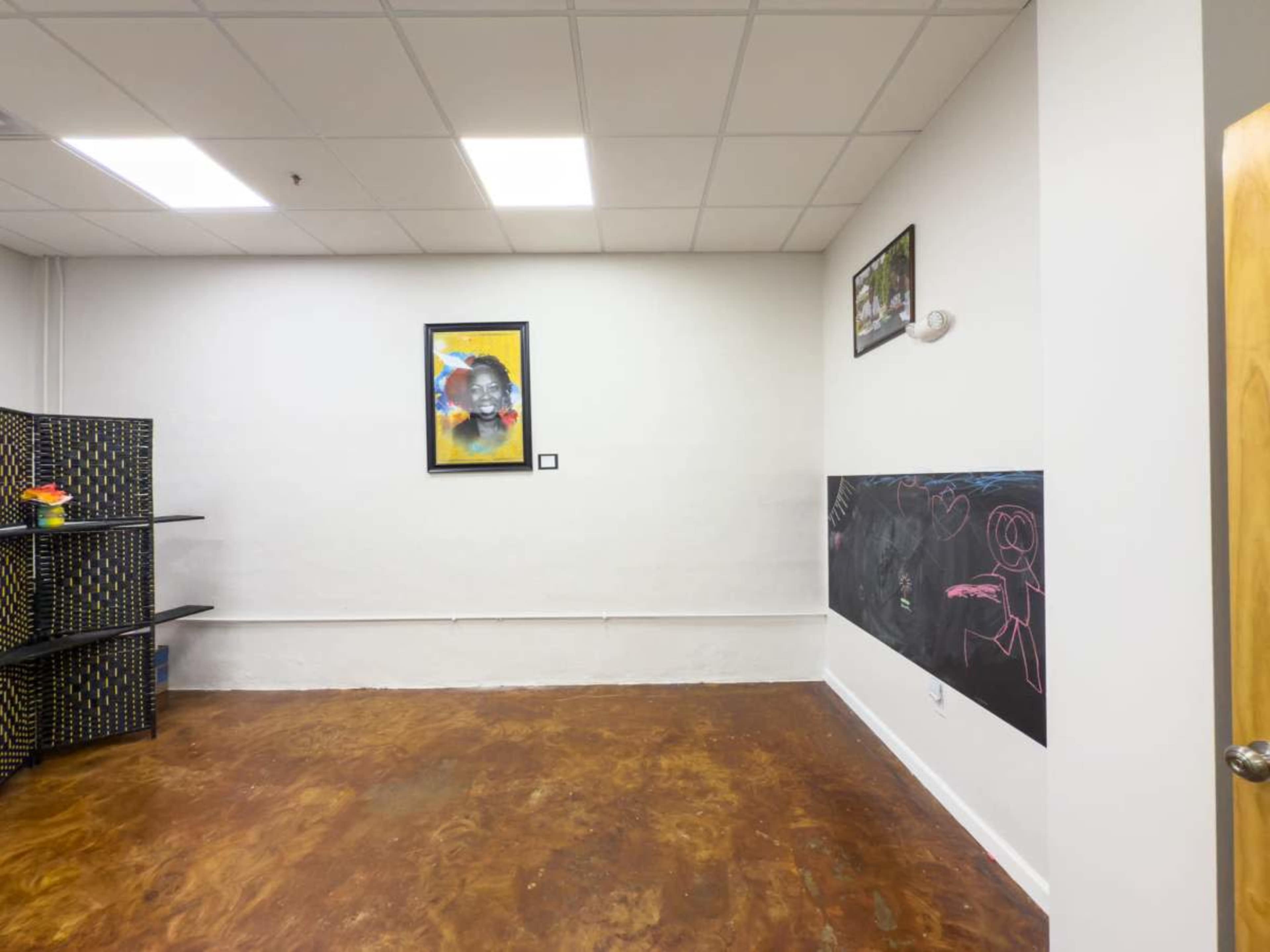 The image depicts a sparsely furnished room with a polished concrete floor, a framed artwork on the wall, and a section of chalkboard on one side.