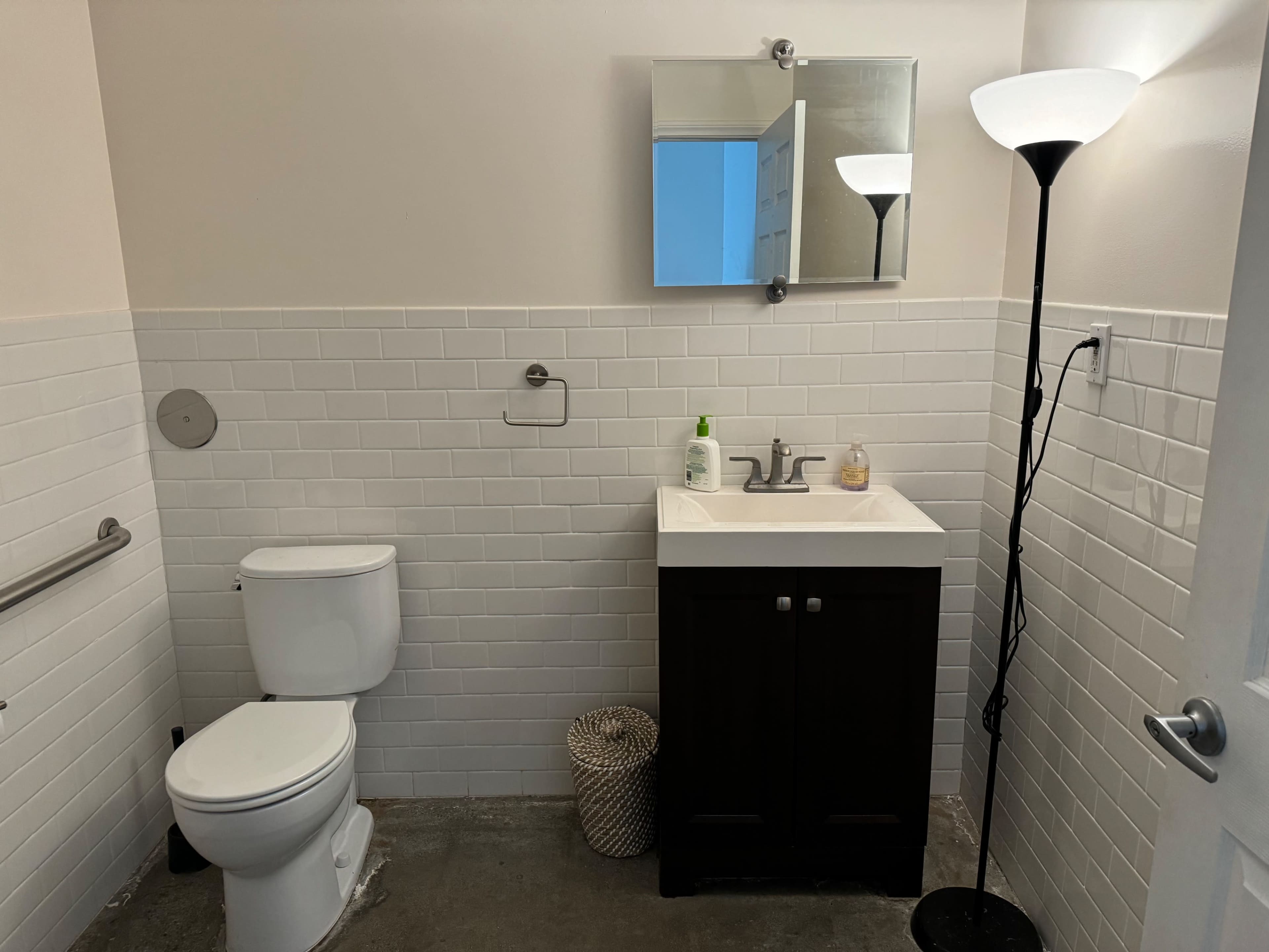 A small bathroom featuring a toilet, a sink with a cabinet below, a mirror above the sink, and a lamp in the corner.