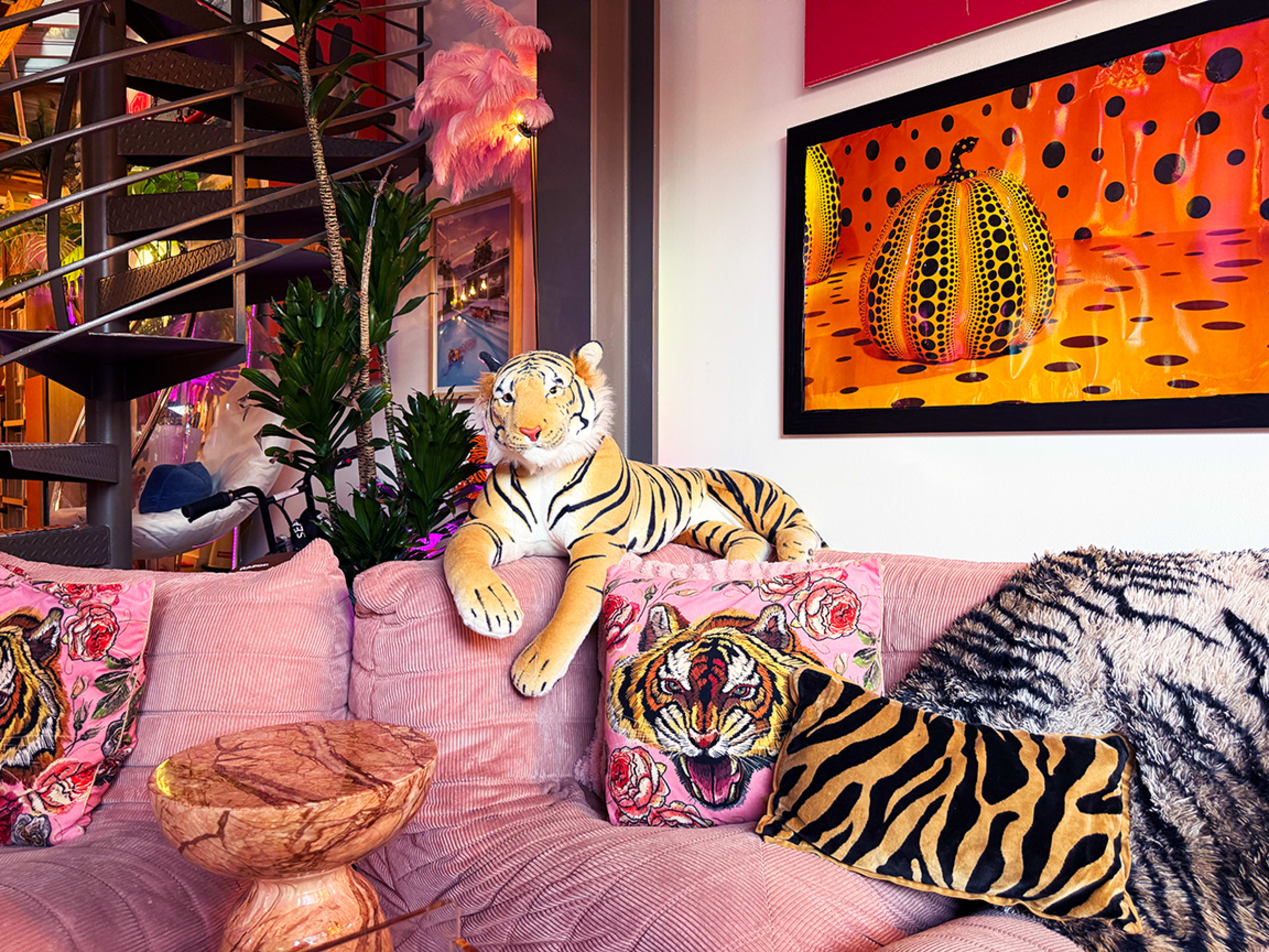 A plush pink sofa with tiger-patterned pillows and a decorative tiger statue is placed next to a colorful artwork featuring polka-dotted pumpkins.