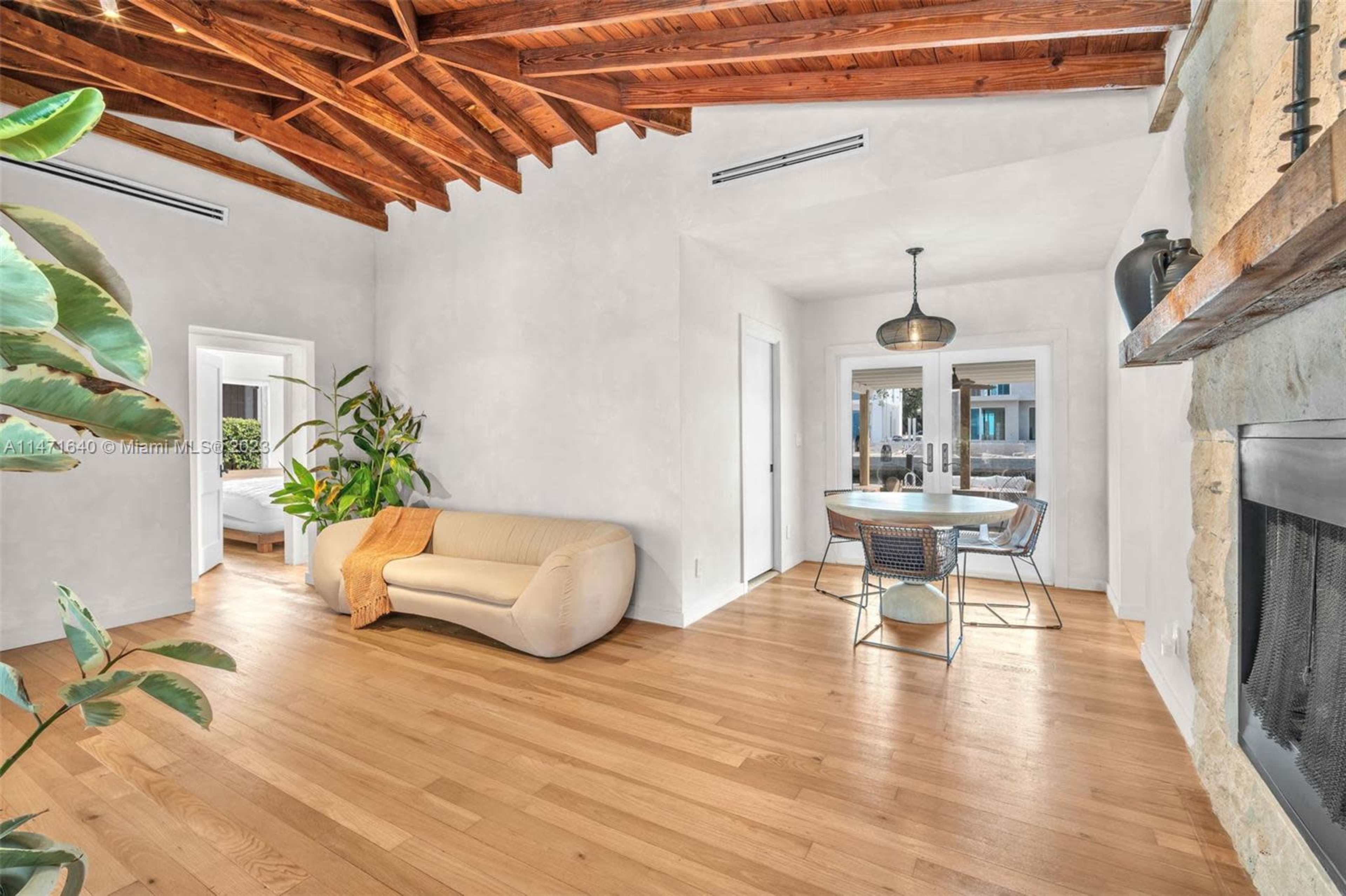 The image shows a modern living room with a light-colored sofa, a round dining table, and exposed wooden beams on the ceiling.