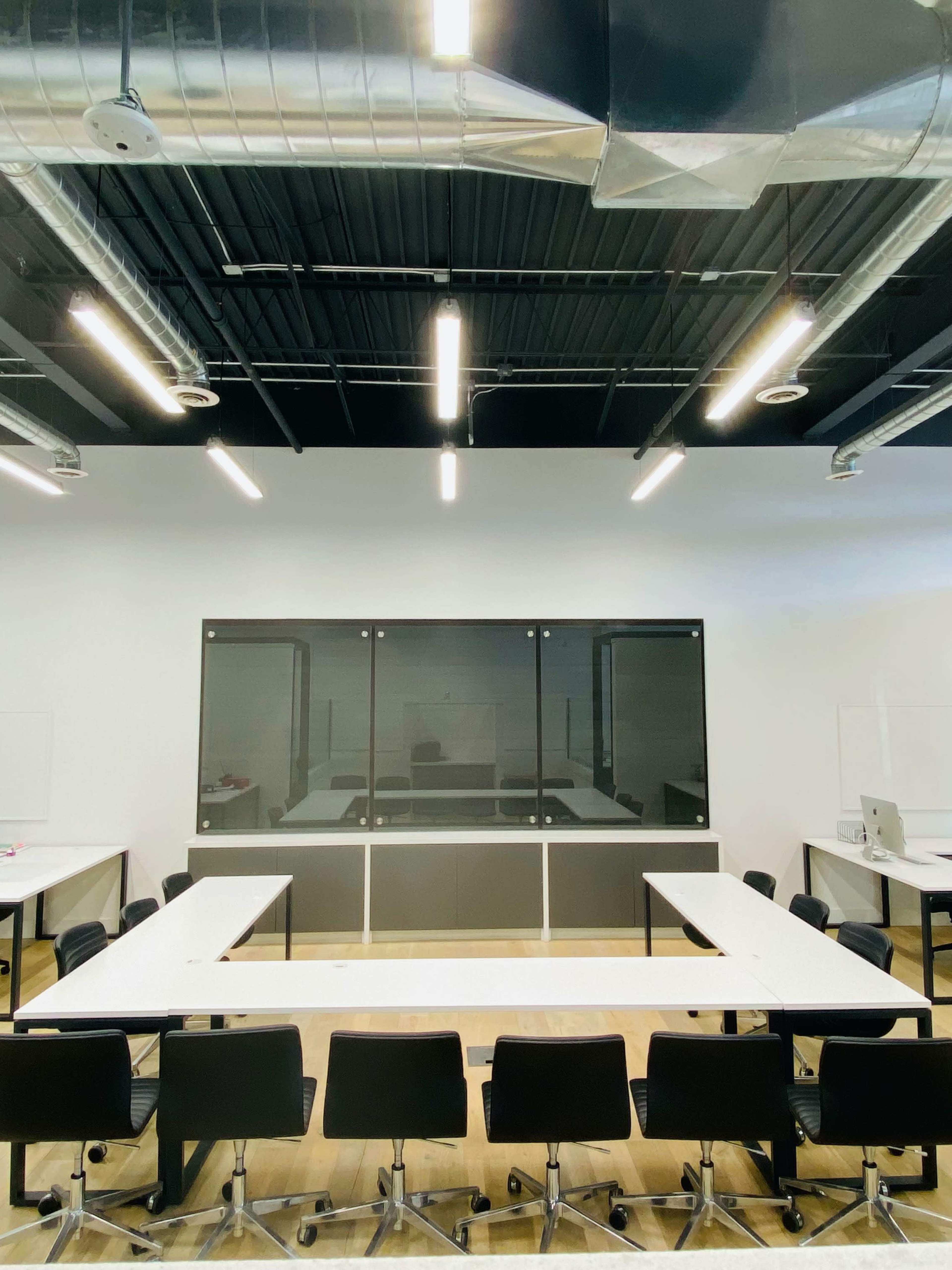 A minimalist conference room features a large rectangular table surrounded by black swivel chairs, with a glass wall at one end.