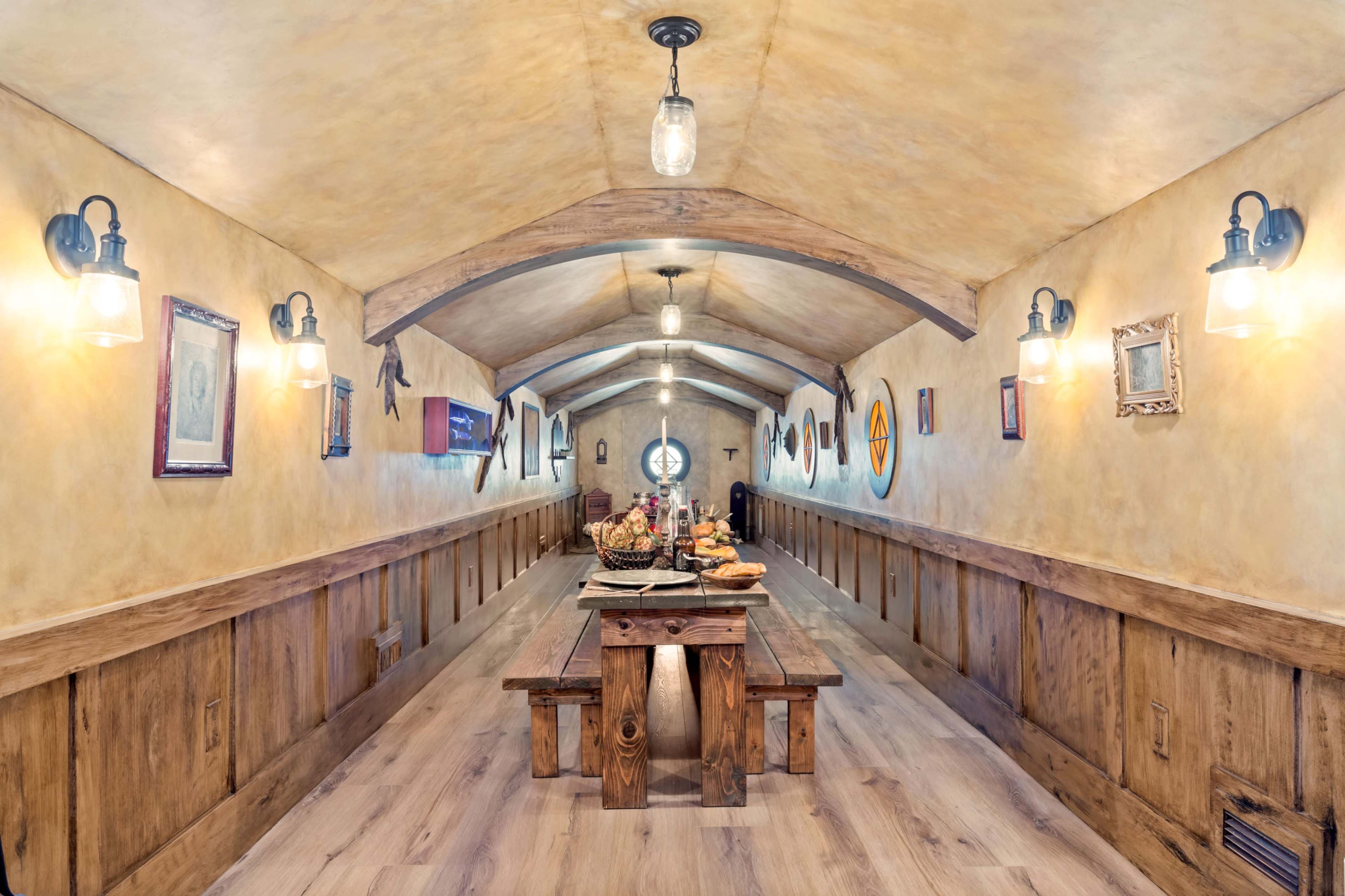 The image shows a long, narrow dining hall with wooden walls, a large table set with food, and various framed artworks lining the walls.