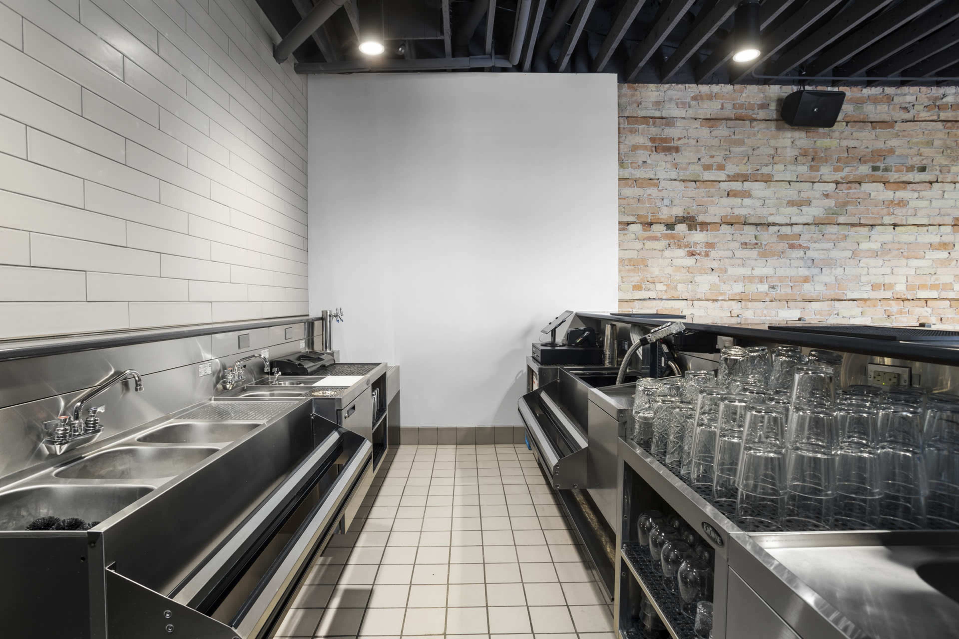 The image shows a commercial kitchen area featuring stainless steel sinks and a glass-washing station against a backdrop of white tiled and exposed brick walls.