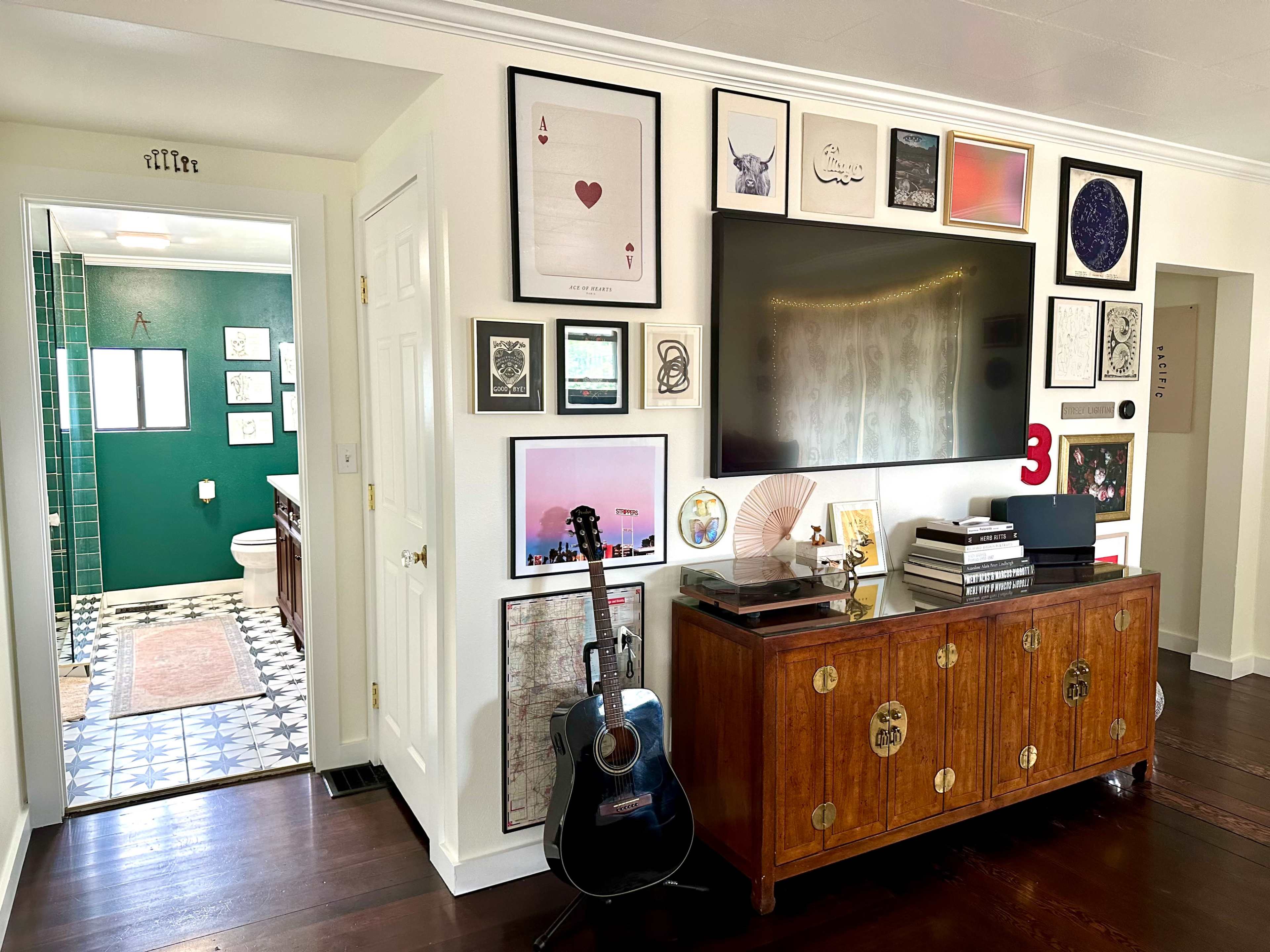 The image shows a modern living room with a wooden cabinet, an acoustic guitar, and a gallery wall of framed artwork, leading to a bathroom with green walls and patterned flooring.
