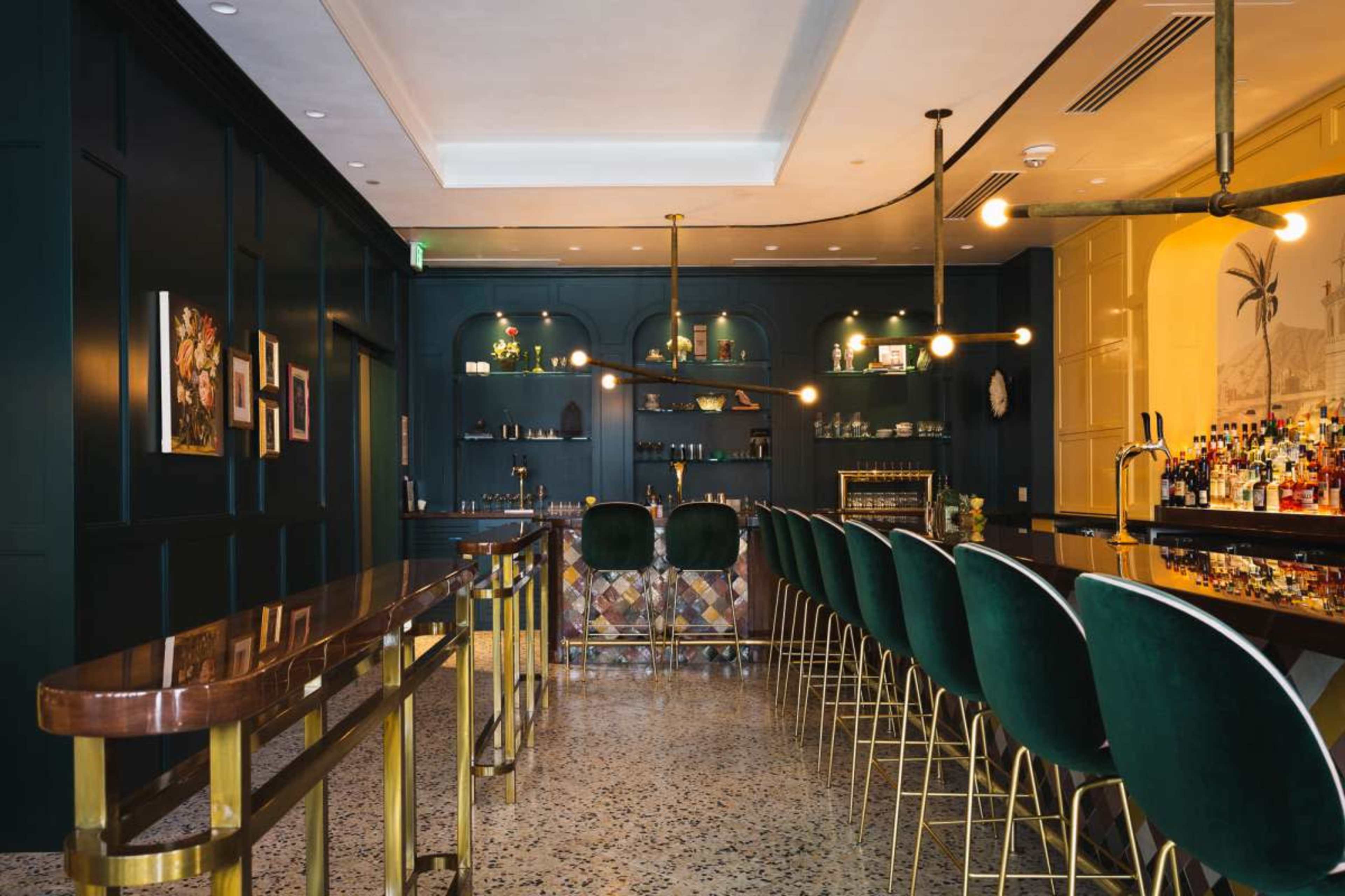 The image shows a stylish bar area featuring golden accents, green velvet bar stools, and a variety of bottles displayed on shelves against a dark blue wall.