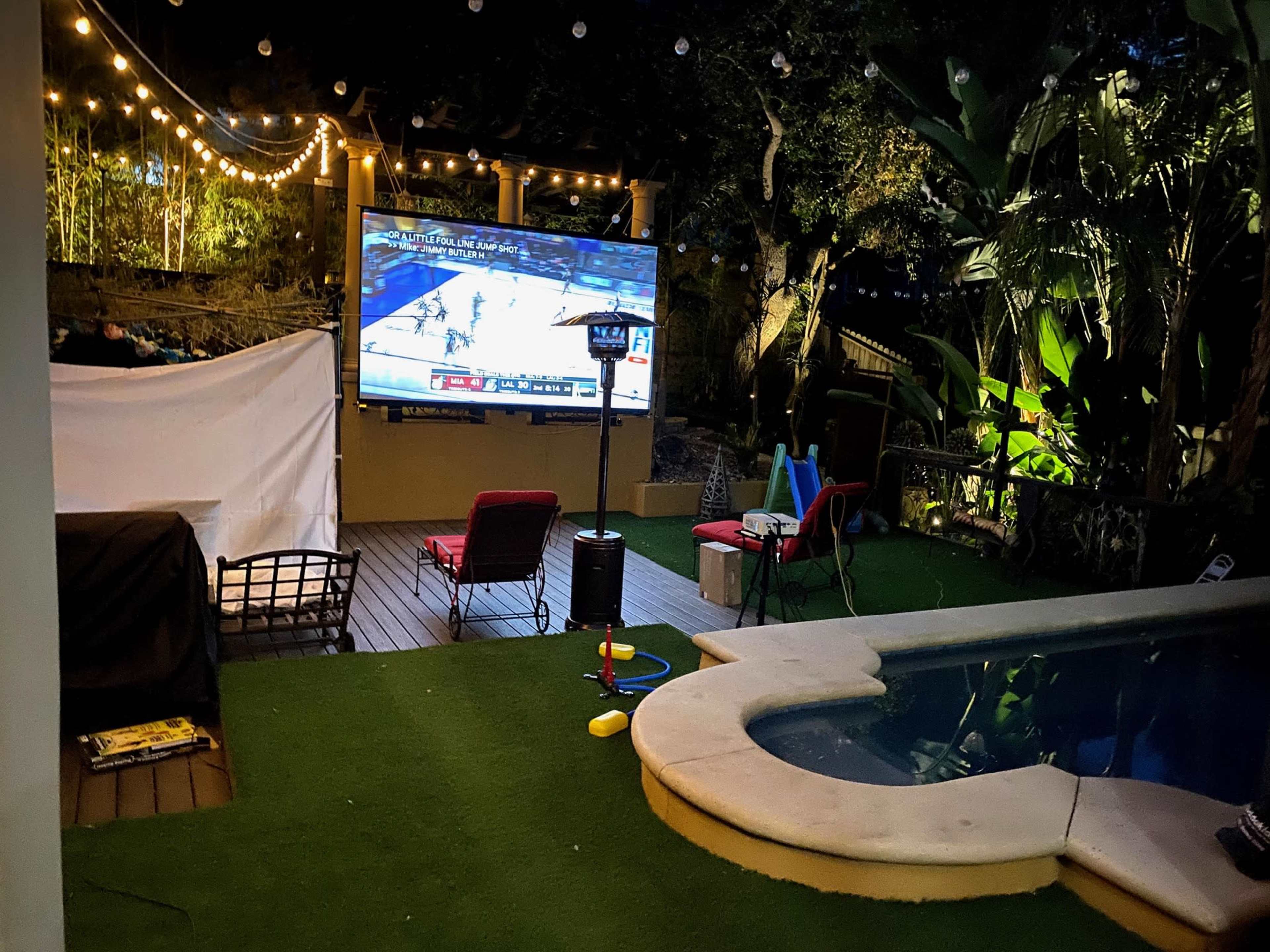 A backyard patio features a large screen displaying a basketball game, surrounded by outdoor seating, string lights, and a pool.