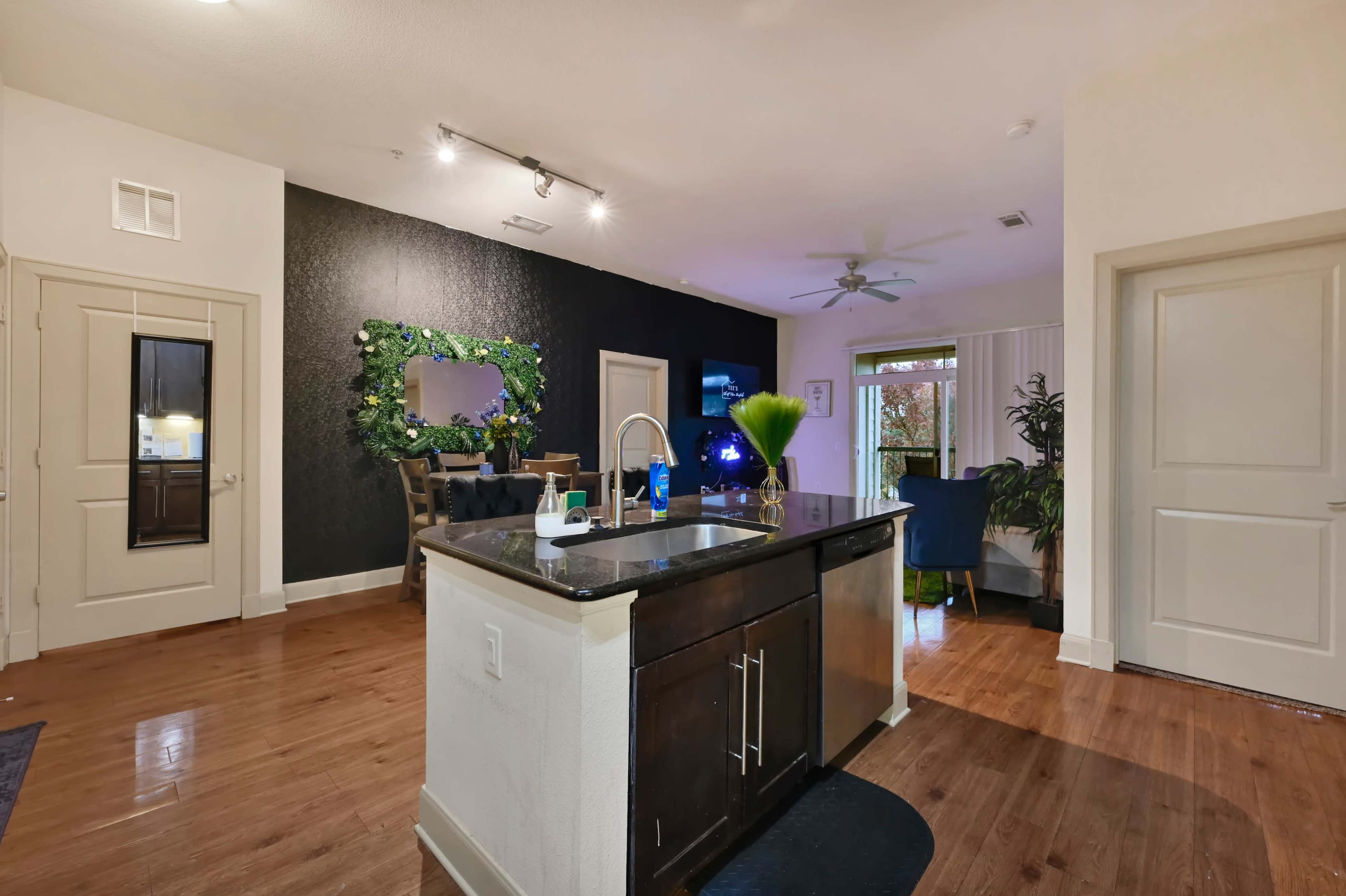 The image shows a modern kitchen and living area with hardwood floors, a dark accent wall, and decorative plants.