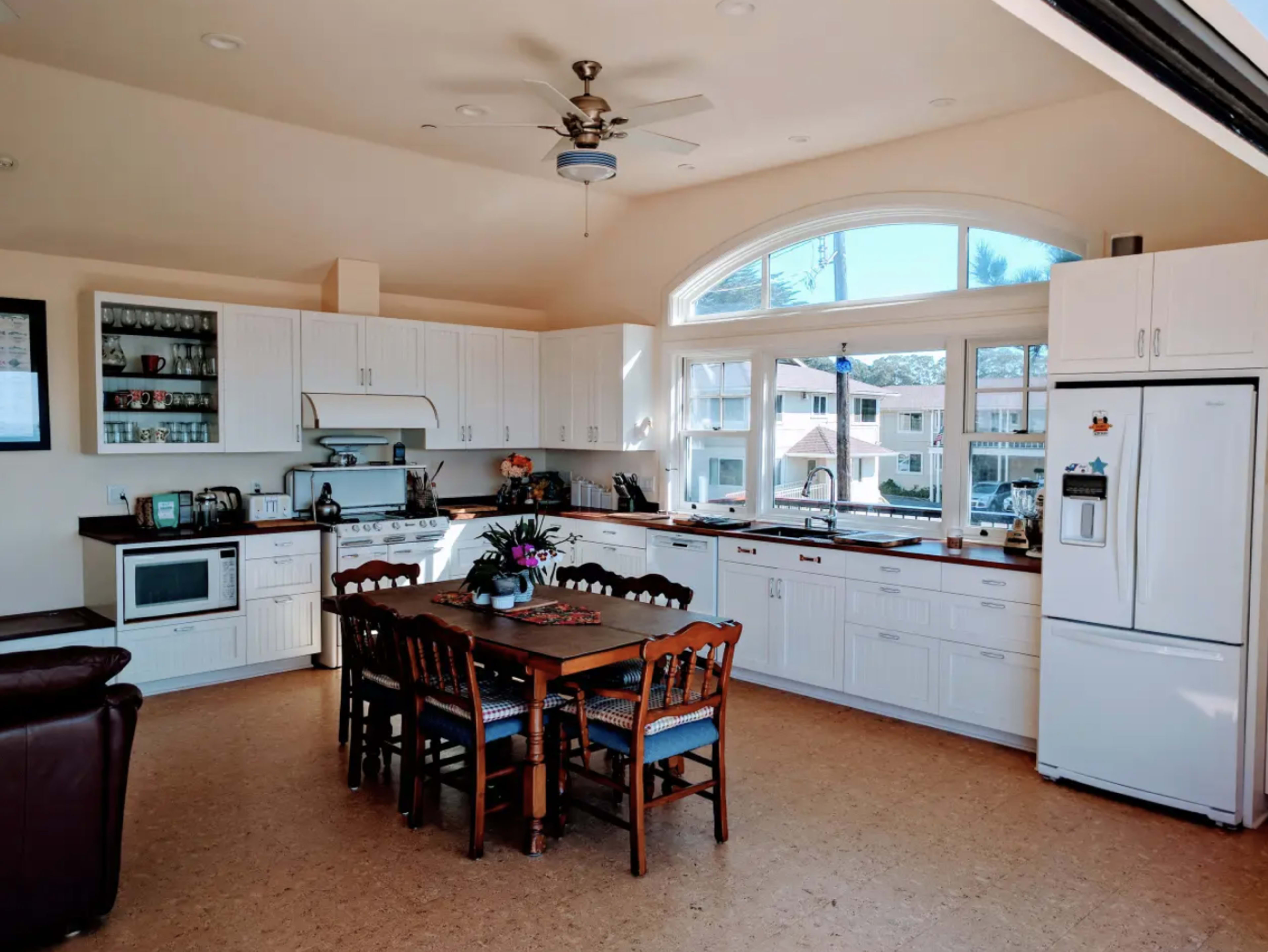 The image shows a spacious kitchen with white cabinets, a central dining table surrounded by wooden chairs, and large windows allowing natural light to enter.