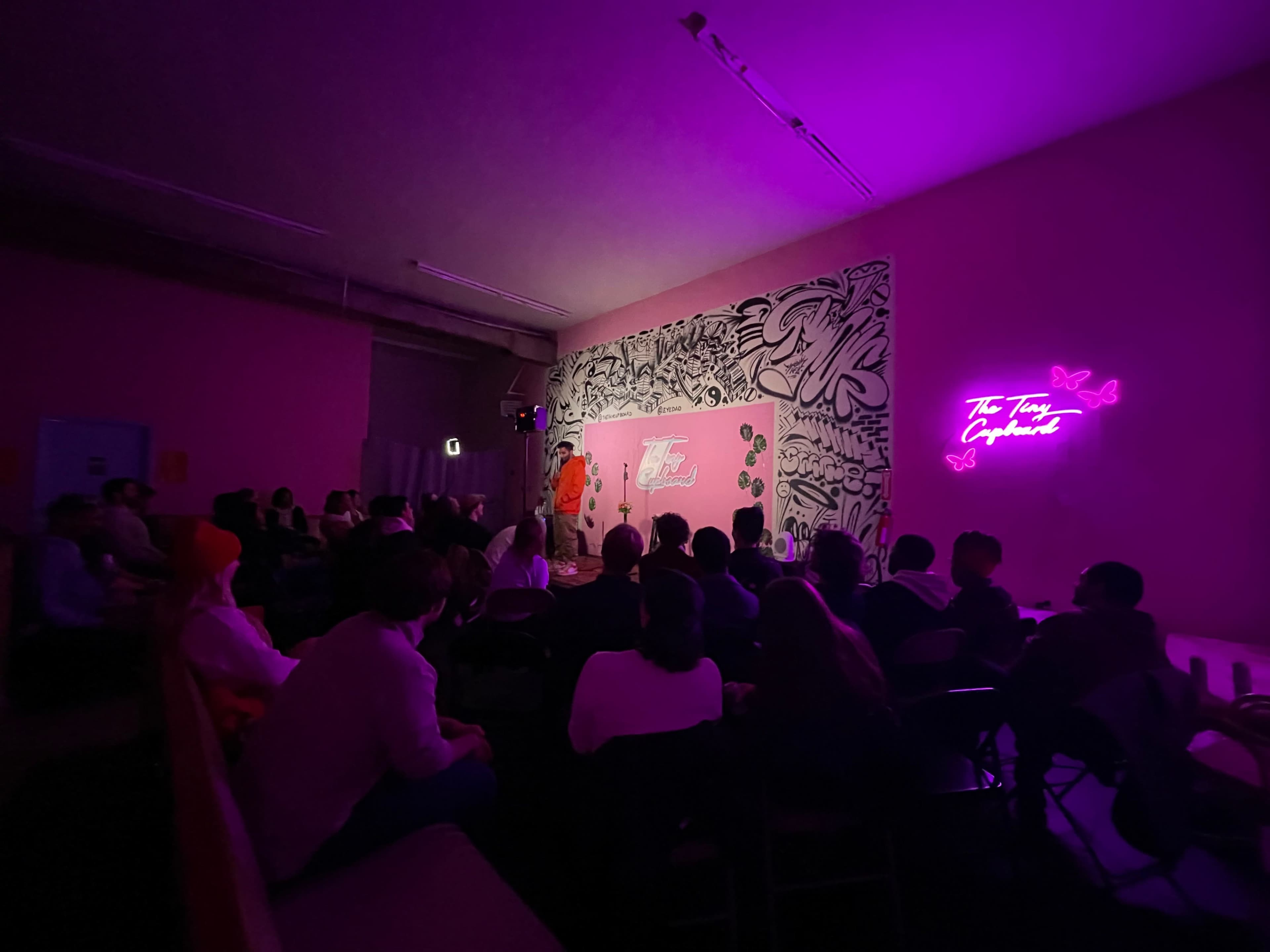 A performer stands on stage in front of an audience in a room with pink walls and vibrant decor, including a neon sign.